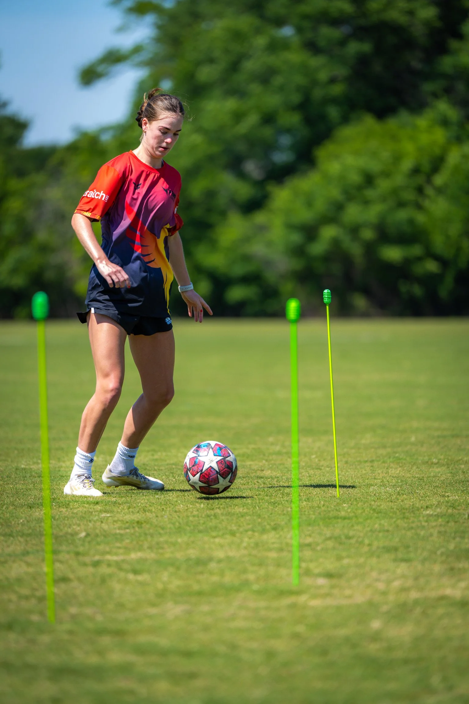 Female soccer player in a colorful jersey practicing dribbling on a grassy field with agility poles.