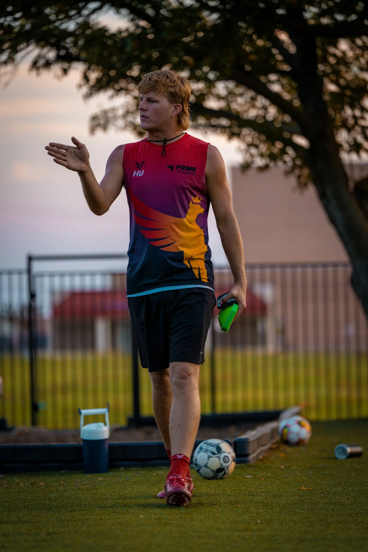 A young man with red hair wearing a red sports tank top, black shorts, and red shoes standing outdoors on a grassy area with soccer balls and water bottles, gesturing with his left hand, during sunset or twilight.
