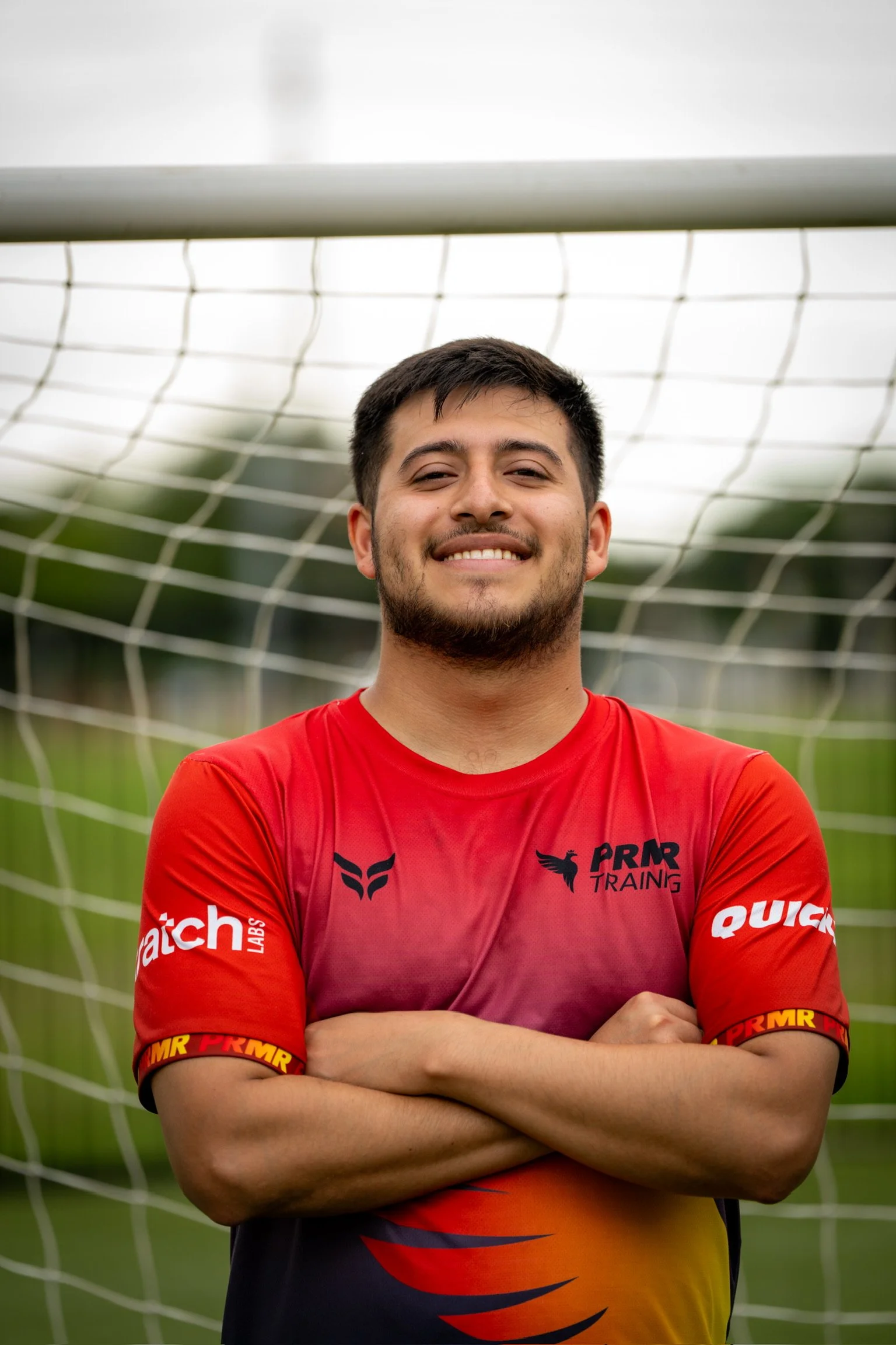 Young man standing in front of a soccer goalpost, smiling, wearing a red and black sports jersey with sponsor logos.