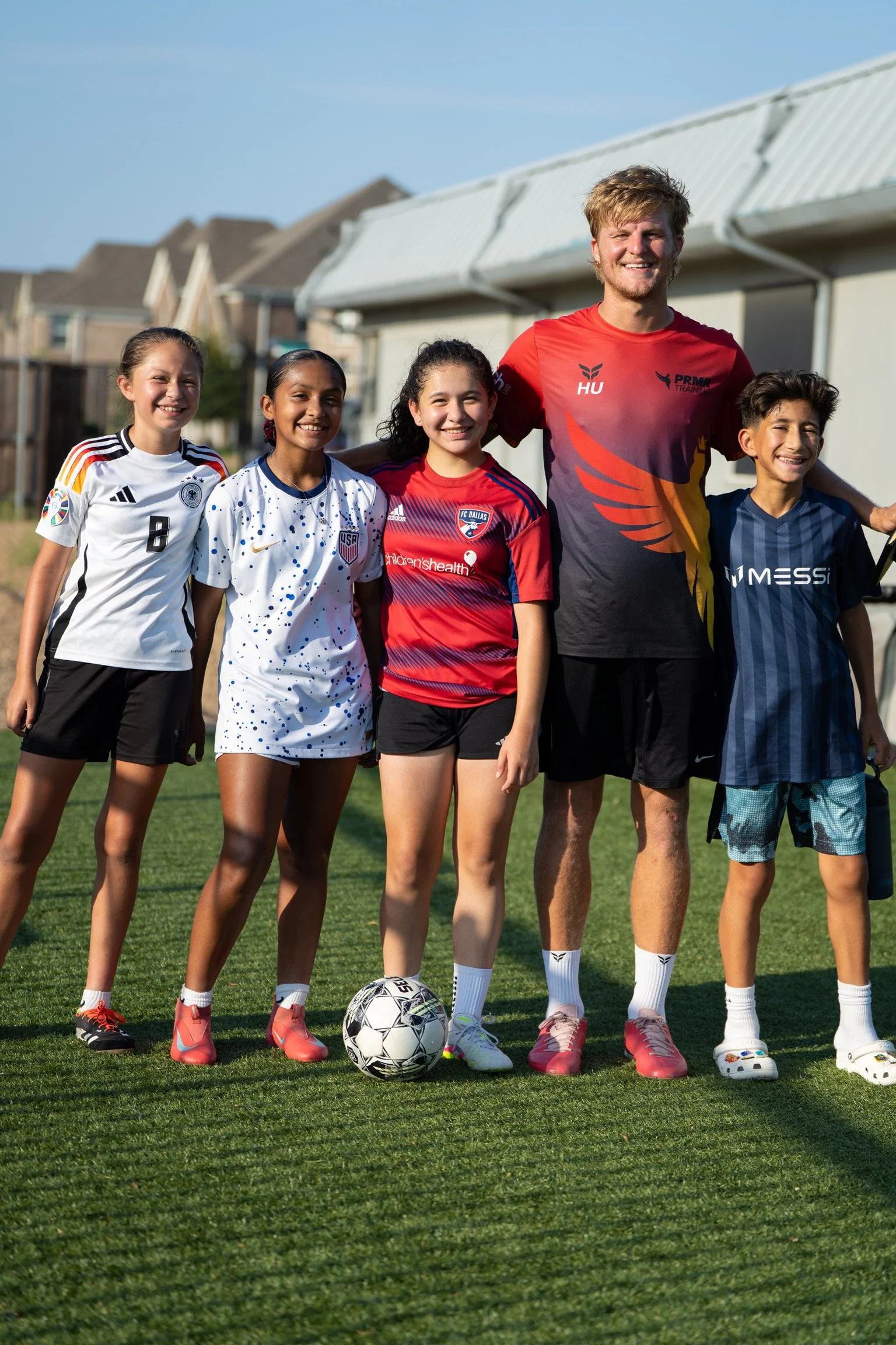 A group of young girls and a coach standing on a soccer field smiling for a photo. The girls are wearing soccer jerseys and shorts, and there is a soccer ball at their feet. The coach is a young man wearing a colorful training shirt and shorts. The background includes houses and a sunny sky.