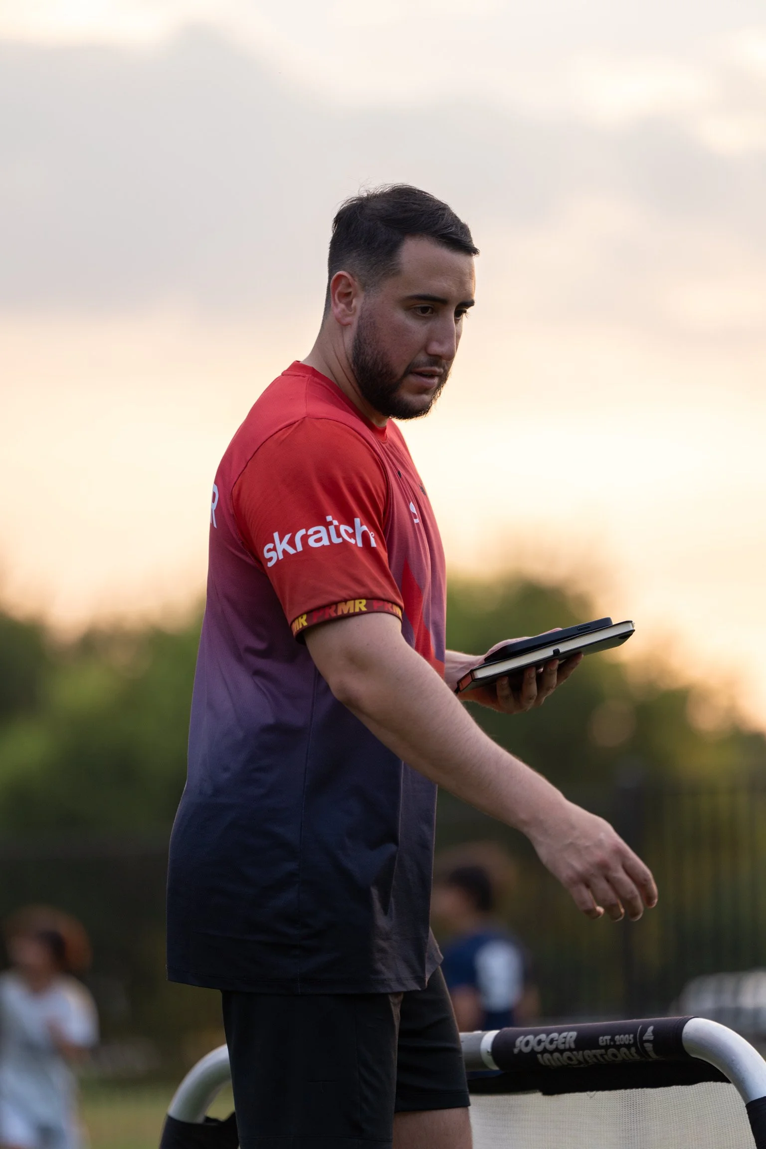 A man with dark hair and beard wearing a red and purple sports shirt stands outdoors during sunset, looking at a device in his hand, with blurred spectators in the background.