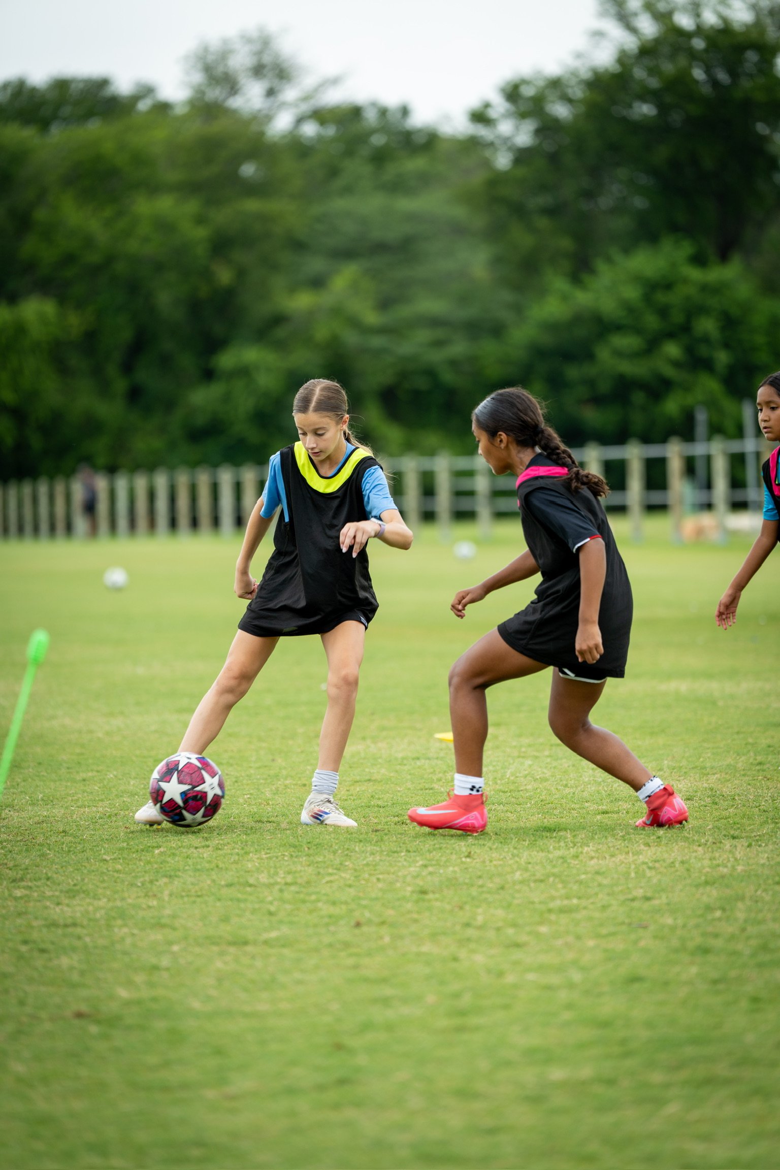 Three young girls playing soccer on a grassy field, with a soccer ball and training cones nearby, surrounded by trees in the background.