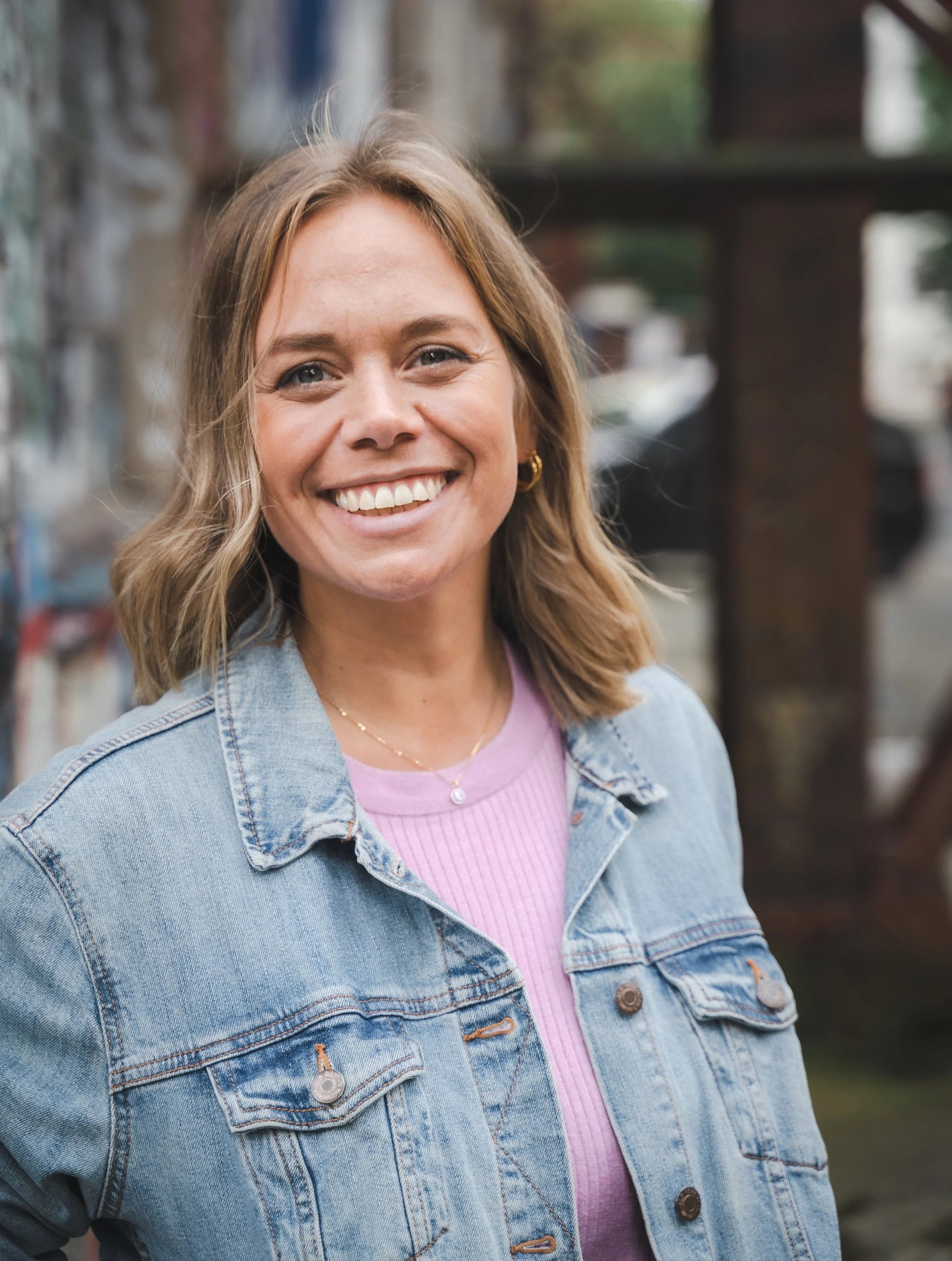 A woman smiling outdoors, wearing a denim jacket and a pink top, with a blurred background of trees and urban surroundings.