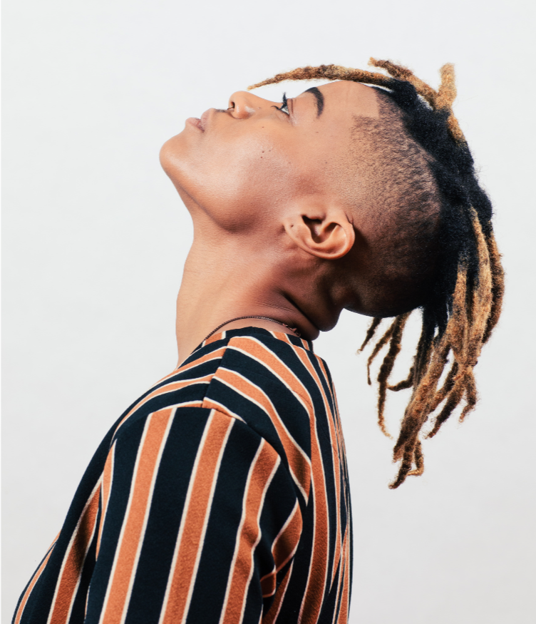 A person with a side profile view showing undercut hairstyle and dreadlocks, wearing a striped shirt against a plain background.
