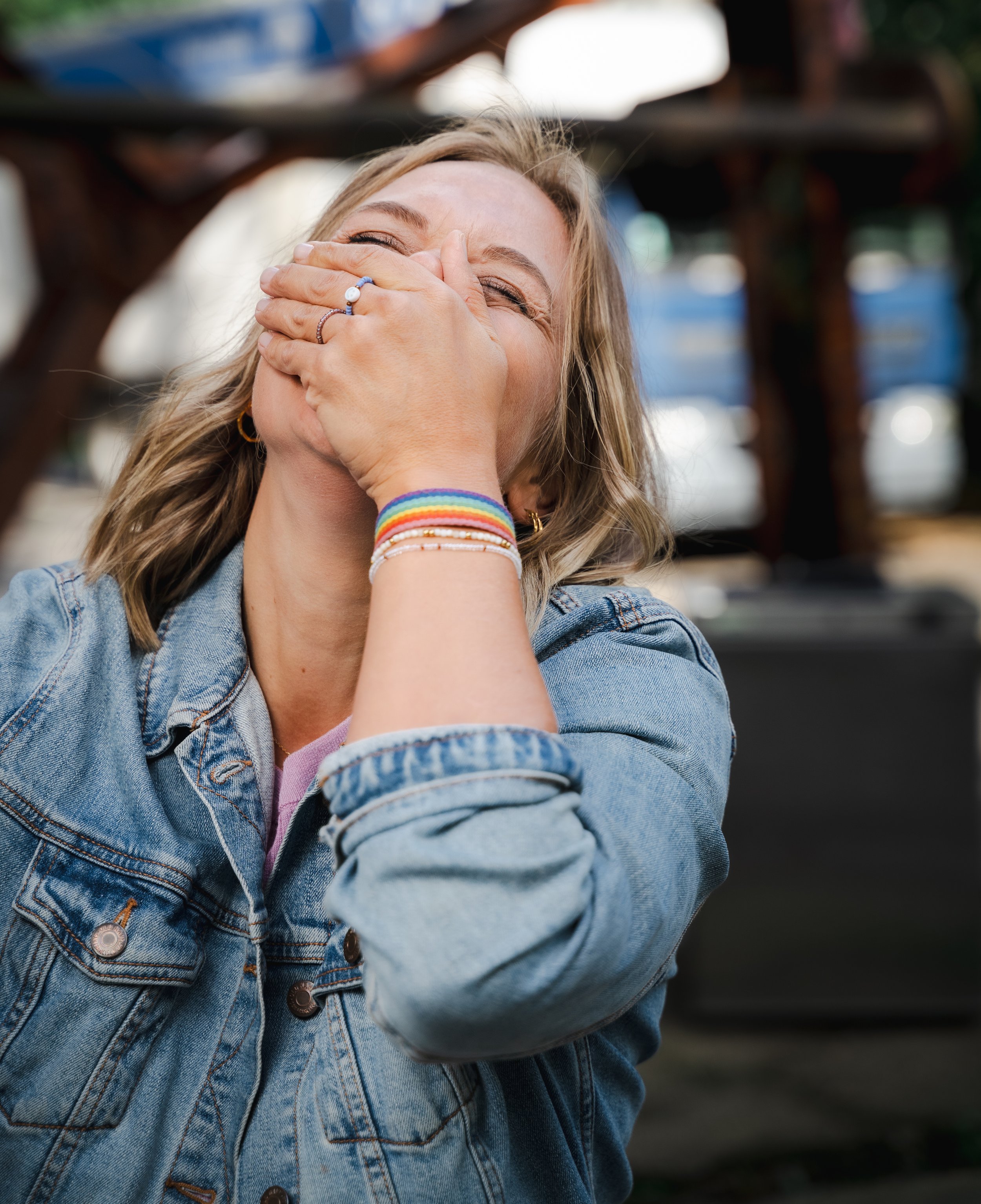 A woman with blonde hair, wearing a denim jacket, laughs and covers her mouth with her hand, which has a ring and rainbow-colored bracelets, in an outdoor setting.