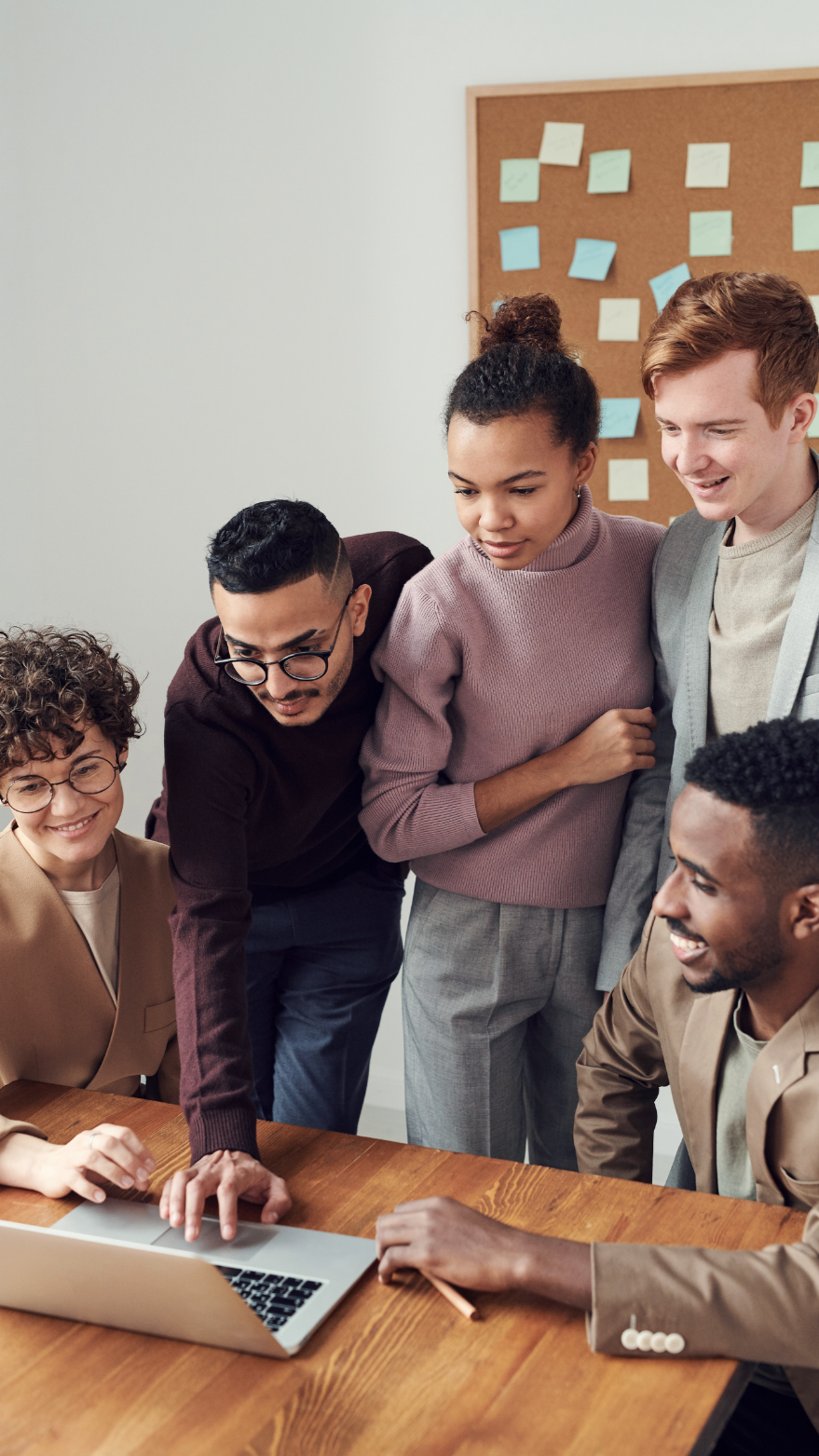 A diverse group of five young professionals gathered around a laptop on a wooden table, looking at the screen with interest during a meeting in an office with a bulletin board in the background.
