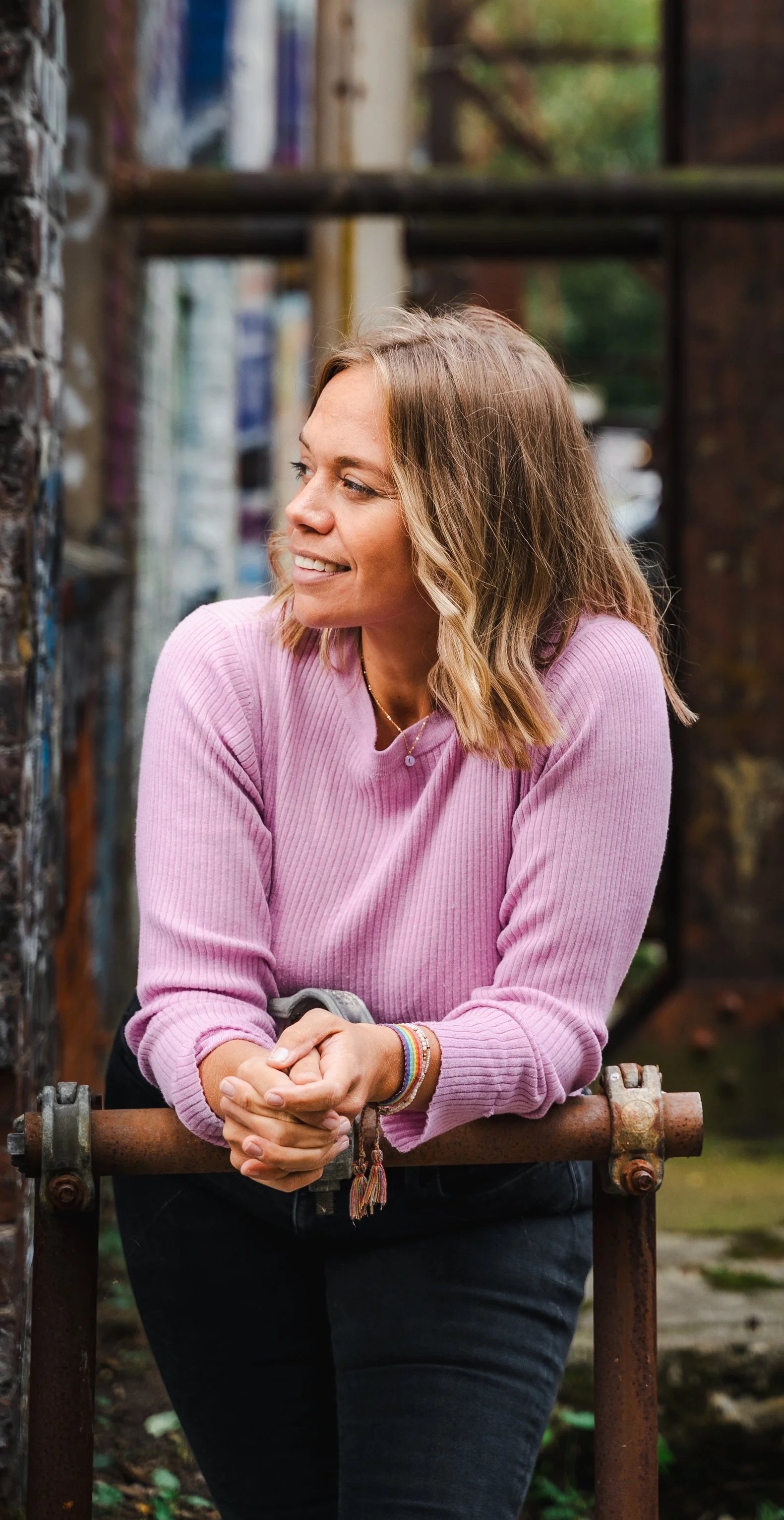 A woman in a pink long-sleeve sweater leaning on a rusty metal railing outdoors, smiling while looking to her left.