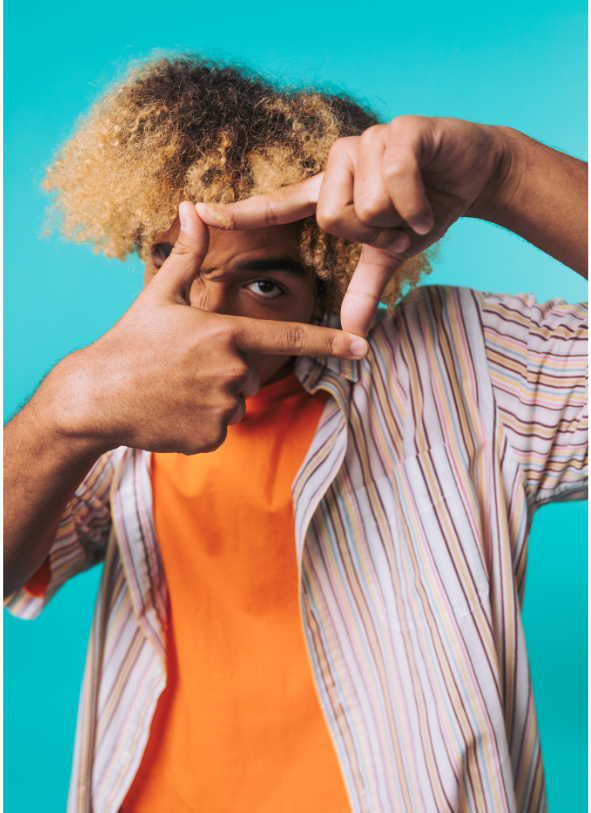 A person with light skin and curly blond hair making a frame with their hands around their eye against a bright blue background, wearing an orange shirt under a striped button-up shirt.