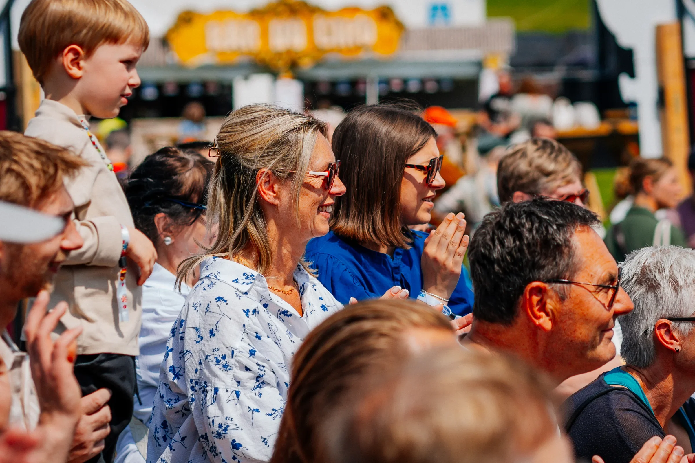 Een groep mensen zit dicht bij elkaar, glimlachend en lachend, buiten op een zonnige dag. Ze lijken te genieten van een evenement of voorstelling.
