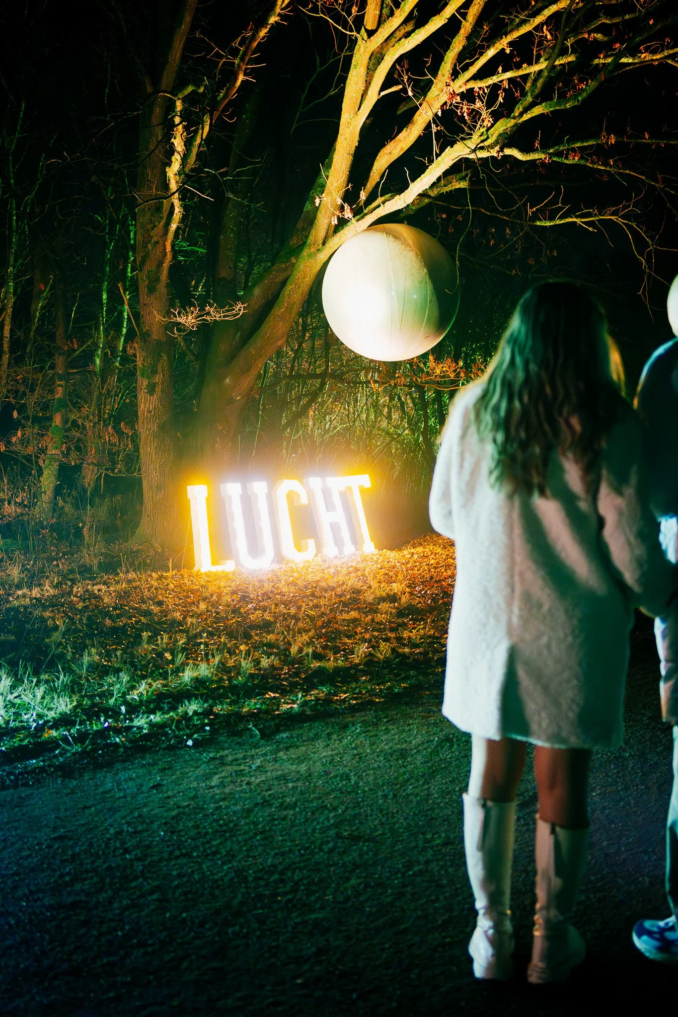 Twee jonge meisjes lopen op een pad in het donker, omringd door bomen met lichtgevende ballonnen en een verlichte tekst 'LUCHT' op de grond.