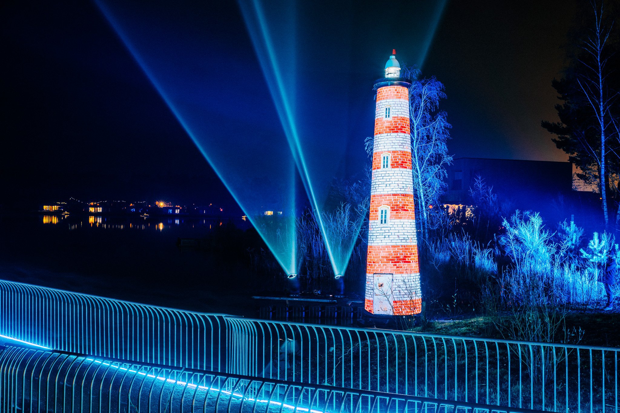 Een verlichte vuurtoren bij nacht met blauwe lichten en waterkant op de voorgrond.
