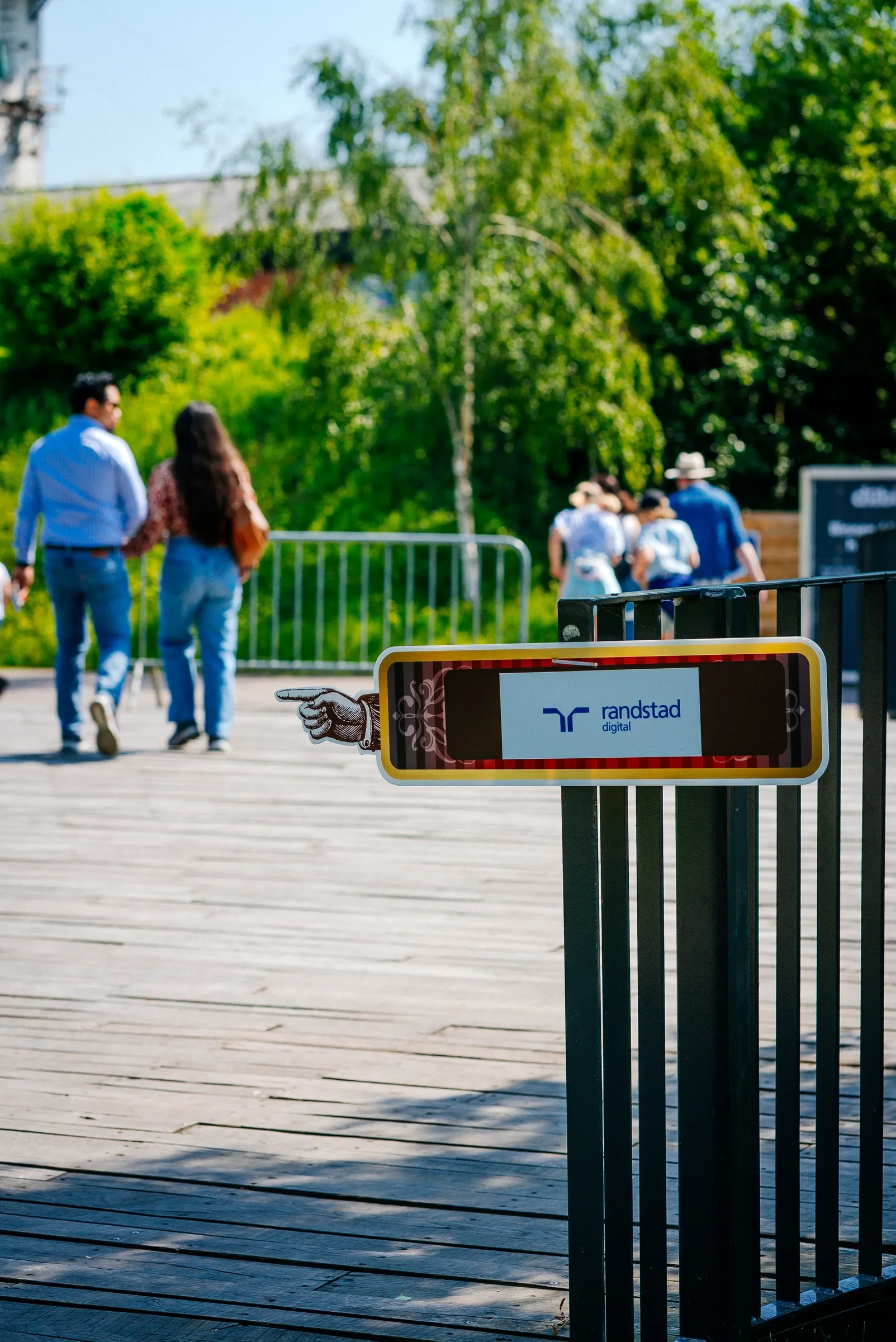 Een bord met het logo van Randstad Digital op een houten promenade, met mensen die op de achtergrond wandelen, omgeven door groene bomen en een blauwe lucht.