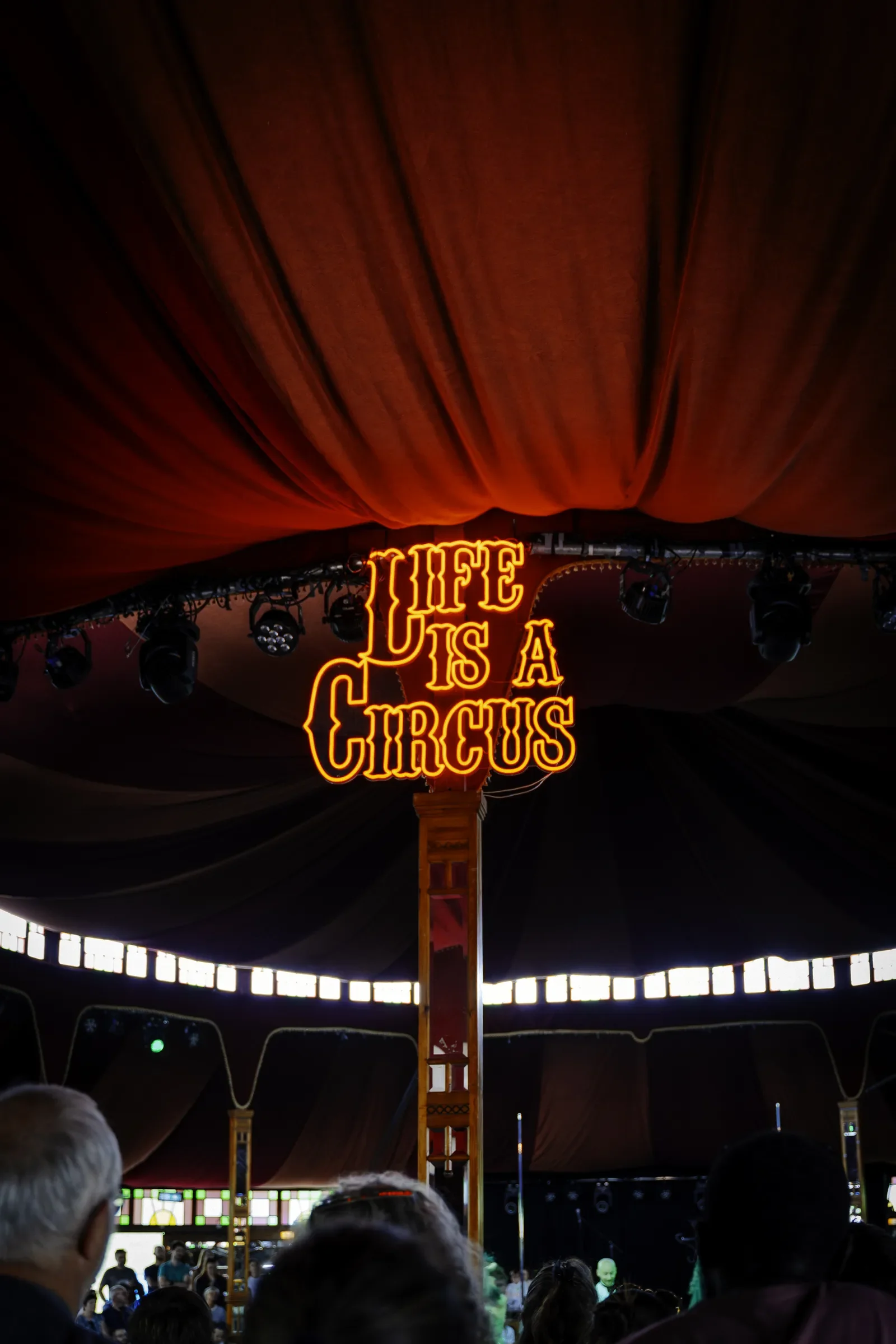 Binnenzijde van een circustent met een neonverlichte bord met de tekst 'Life is a Circus', omringd door mensen en circustrappen.