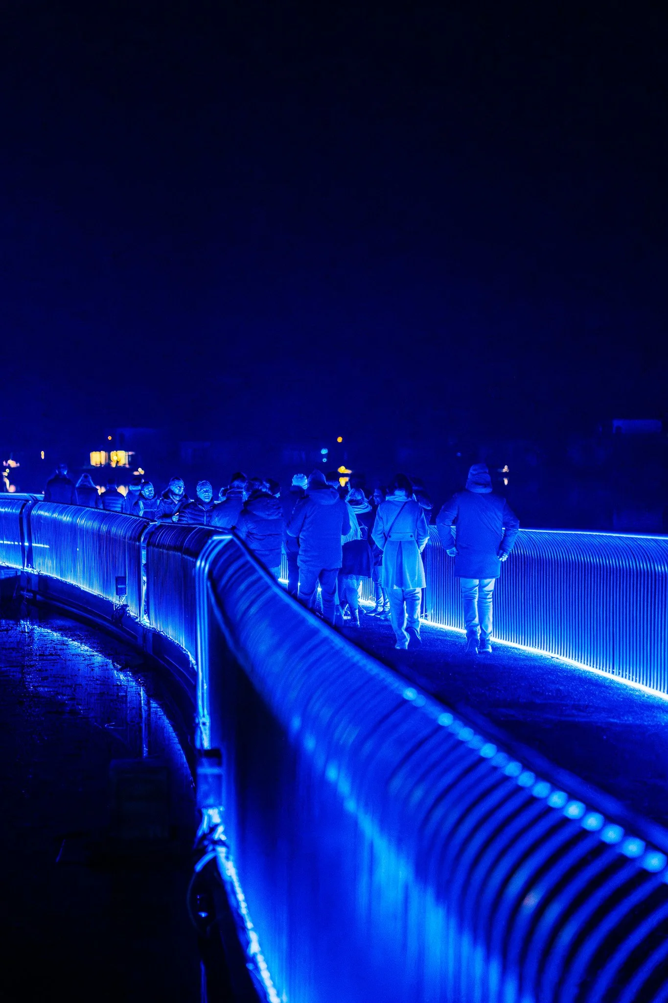 Mensengroep wandelt over een verlichte brug 's nachts, omgeven door een waterpartij en verlichte huizen op de achtergrond.