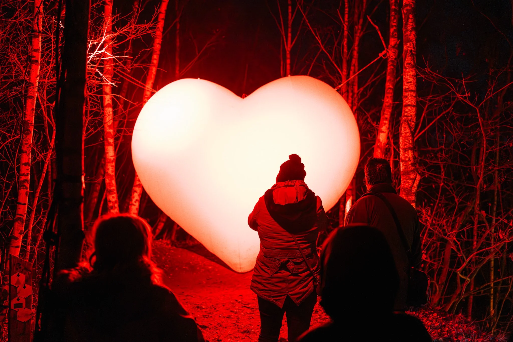 Een groep mensen die naar een grote verlichte hartvormige ballon kijken in een bos bij nacht, allemaal gekleed in winterkleding.