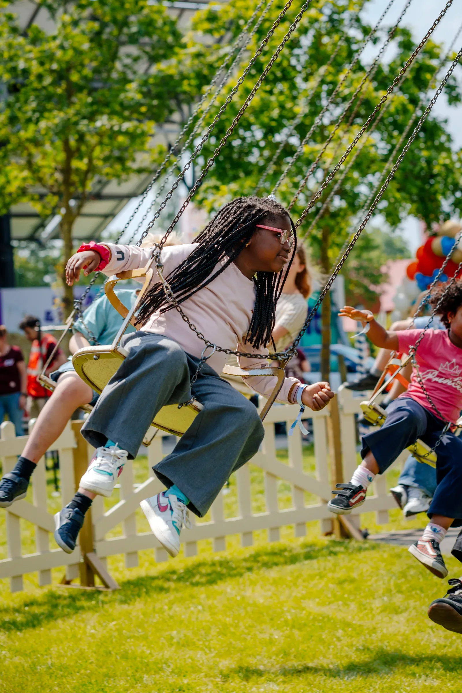 Kinderen op een vrolijk dag in een attractiepark, zittend op een schommel met kettingen, omgeven door groene bomen en een hek in de achtergrond