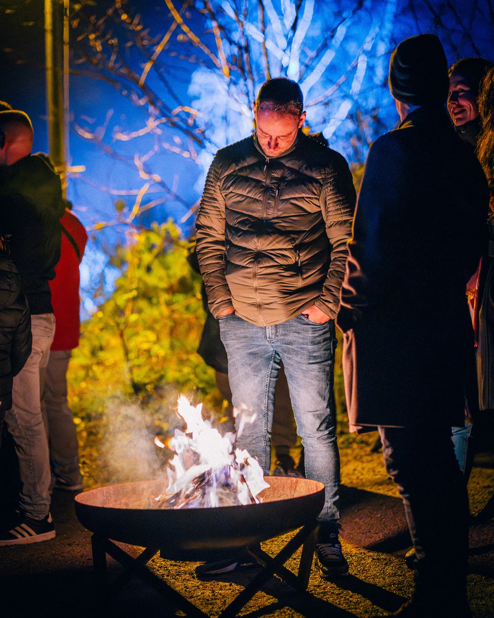 Mensen staan rond een open vuur in de buitenlucht 's avonds, onder een lichtblauw verlichte boom, en genieten van elkaars gezelschap.