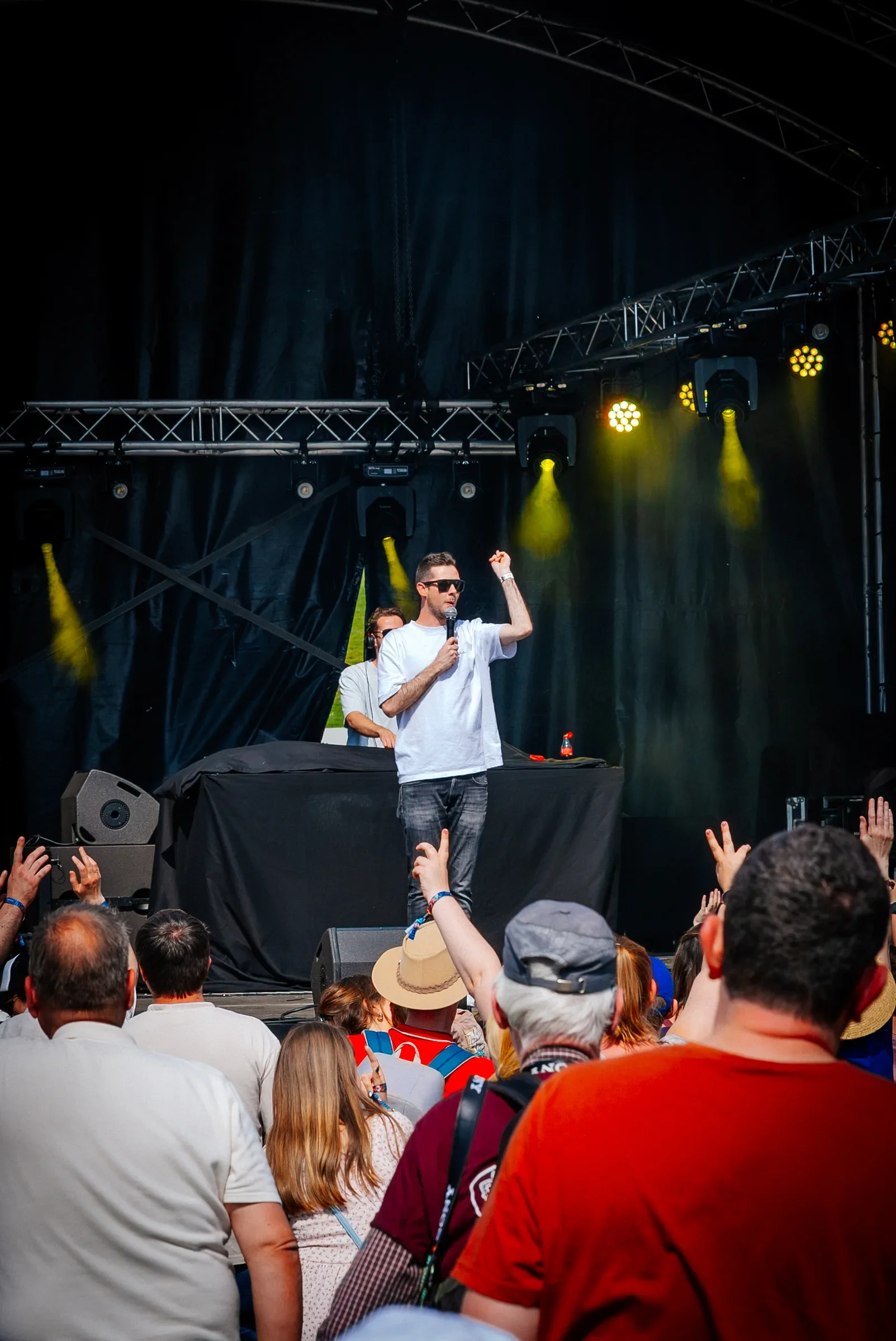 Een persoon op een podium spreekt tot een groot publiek tijdens een concert of evenement, met mensen die omhoog hun handen hebben.