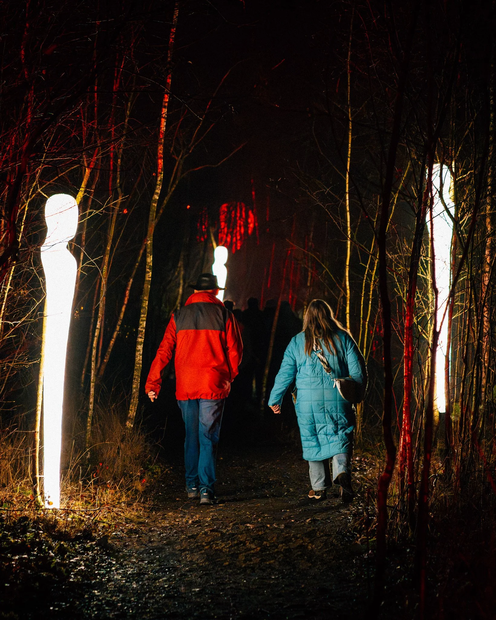 Mensen lopen hand in hand door een bos in de nacht, verlicht door inwaartse lampen en rode lichten in de verte.