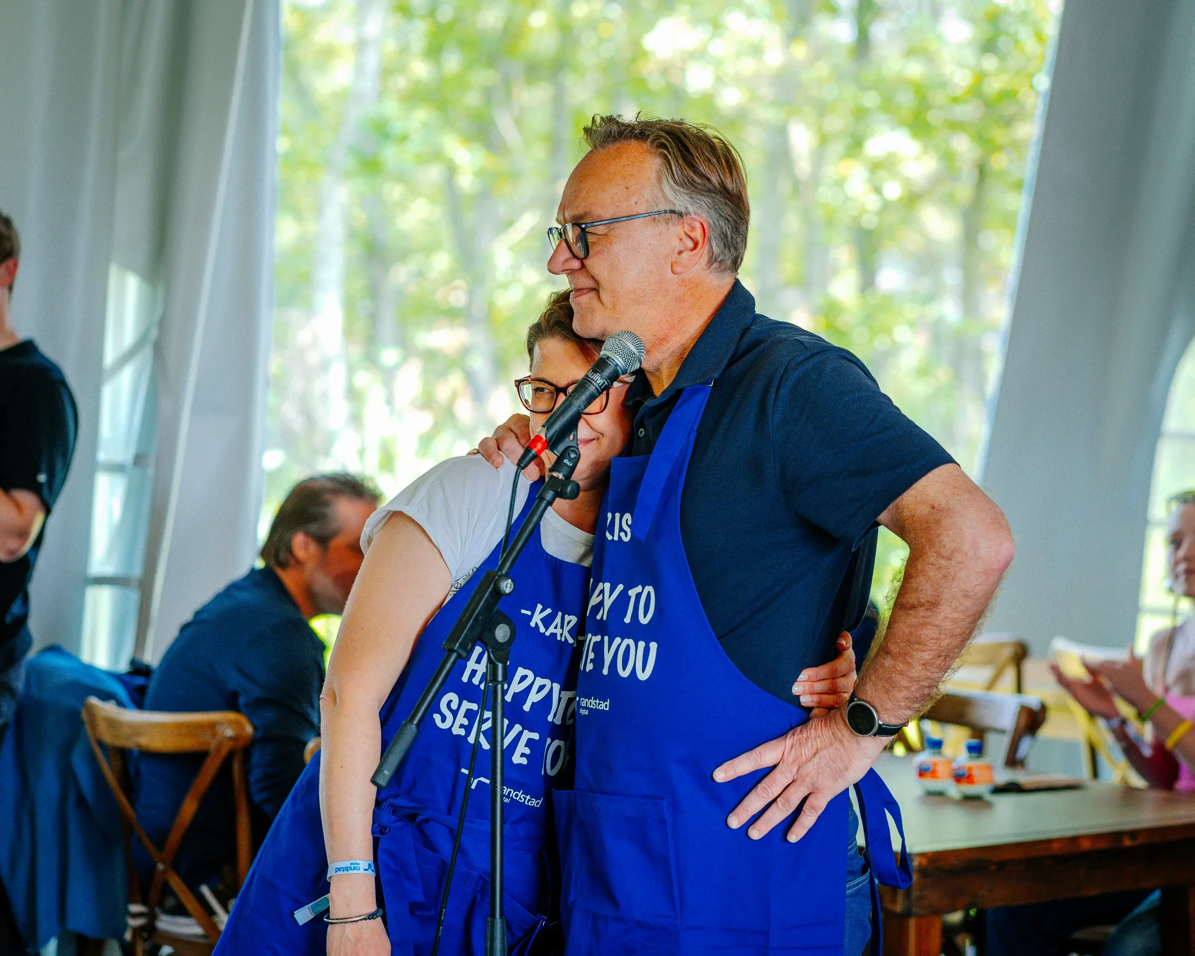 Mannen met bril en grijs haar houdt een microfoon vast en staat dicht bij een vrouw, beiden lachen. Ze dragen blauwe schorten met witte tekst. Ze zijn binnen, met grote ramen en groene bomen buiten.