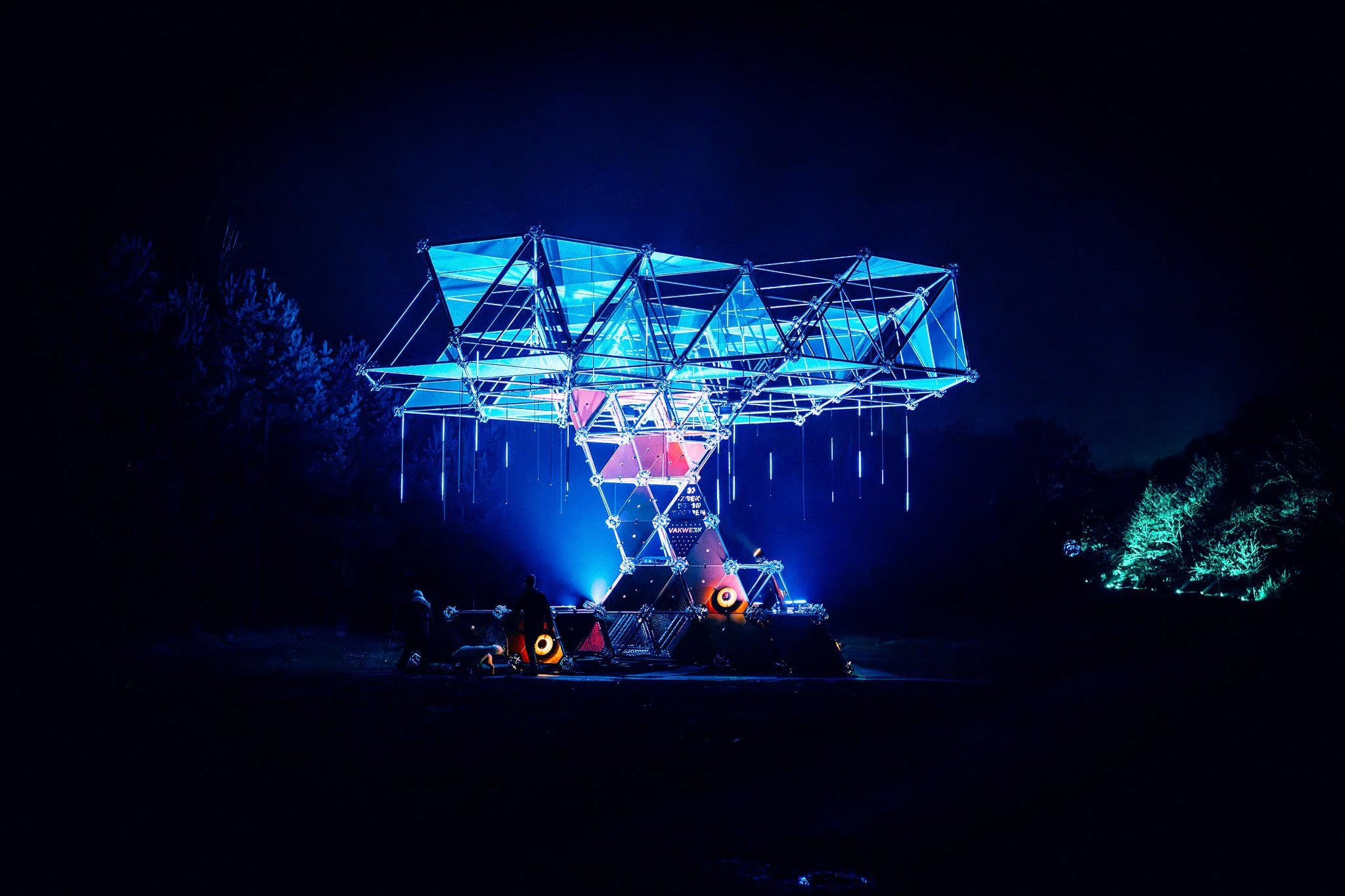 Een verlichte, futuristische structuur met blauwe neonlichten op een donkere achtergrond, mensen staan eromheen.