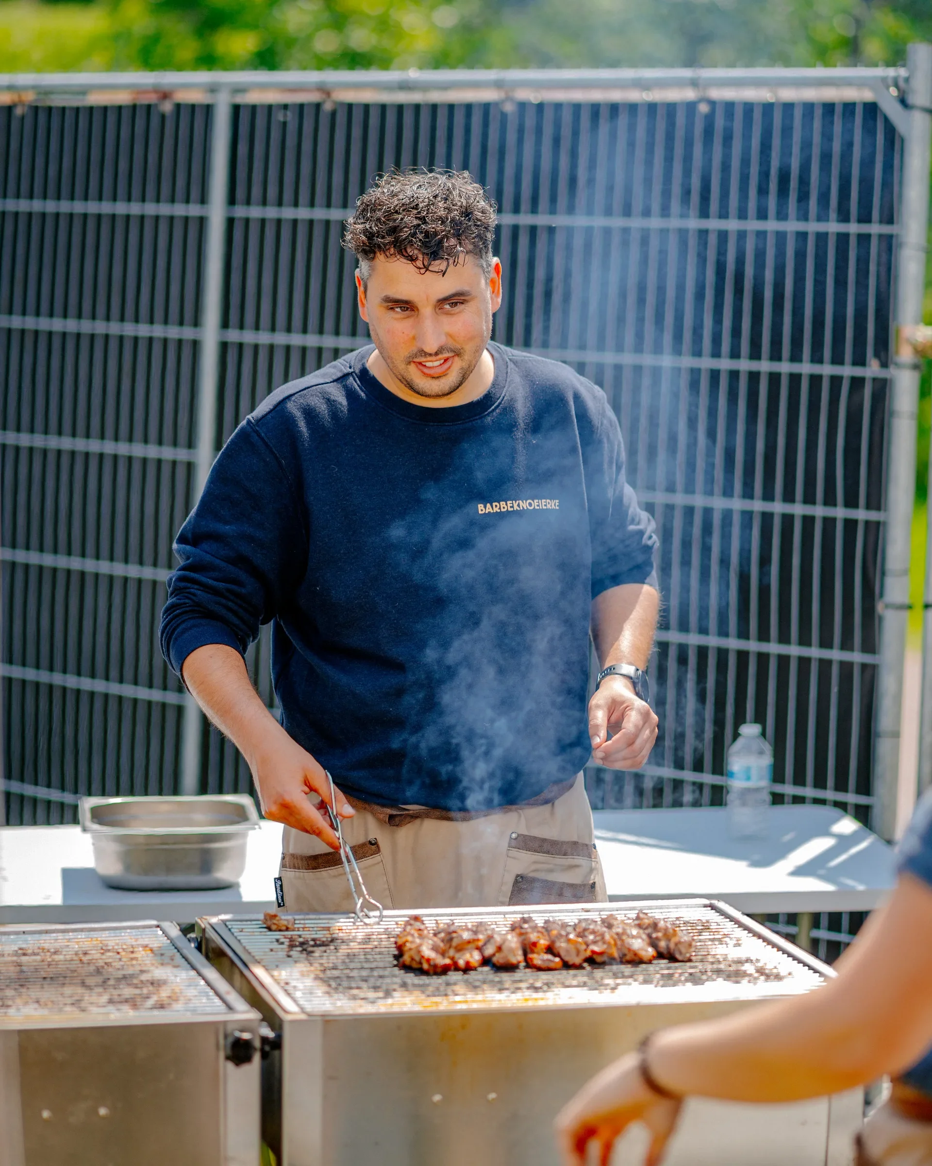 Man grillt vlees op een barbecue buiten op een warme dag, met een metalen hek op de achtergrond.