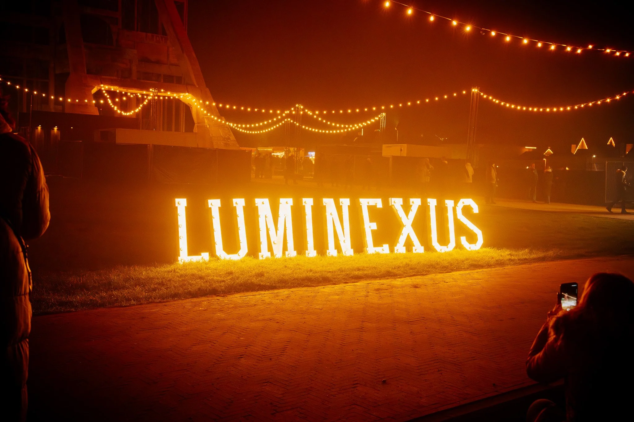 Verlichte 'LUMINEUX' letters bij nacht, met hangende lichtslingers erboven en mensen op de achtergrond.