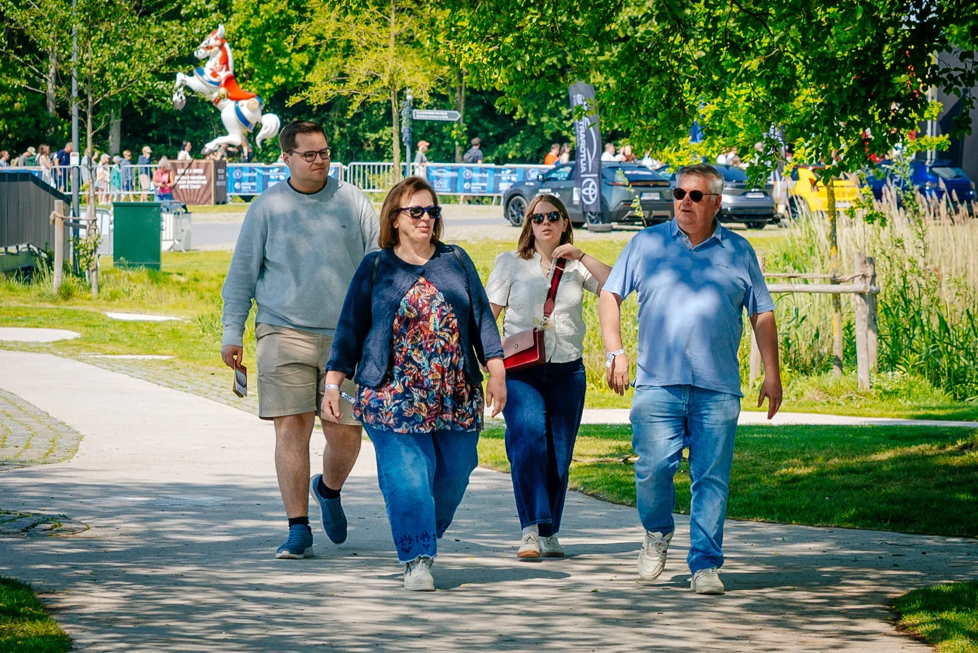 Vijf mensen wandelen op een pad in een park, met bomen en gras rondom. Er zijn meer mensen op de achtergrond en een standbeeld van een sierpaard.