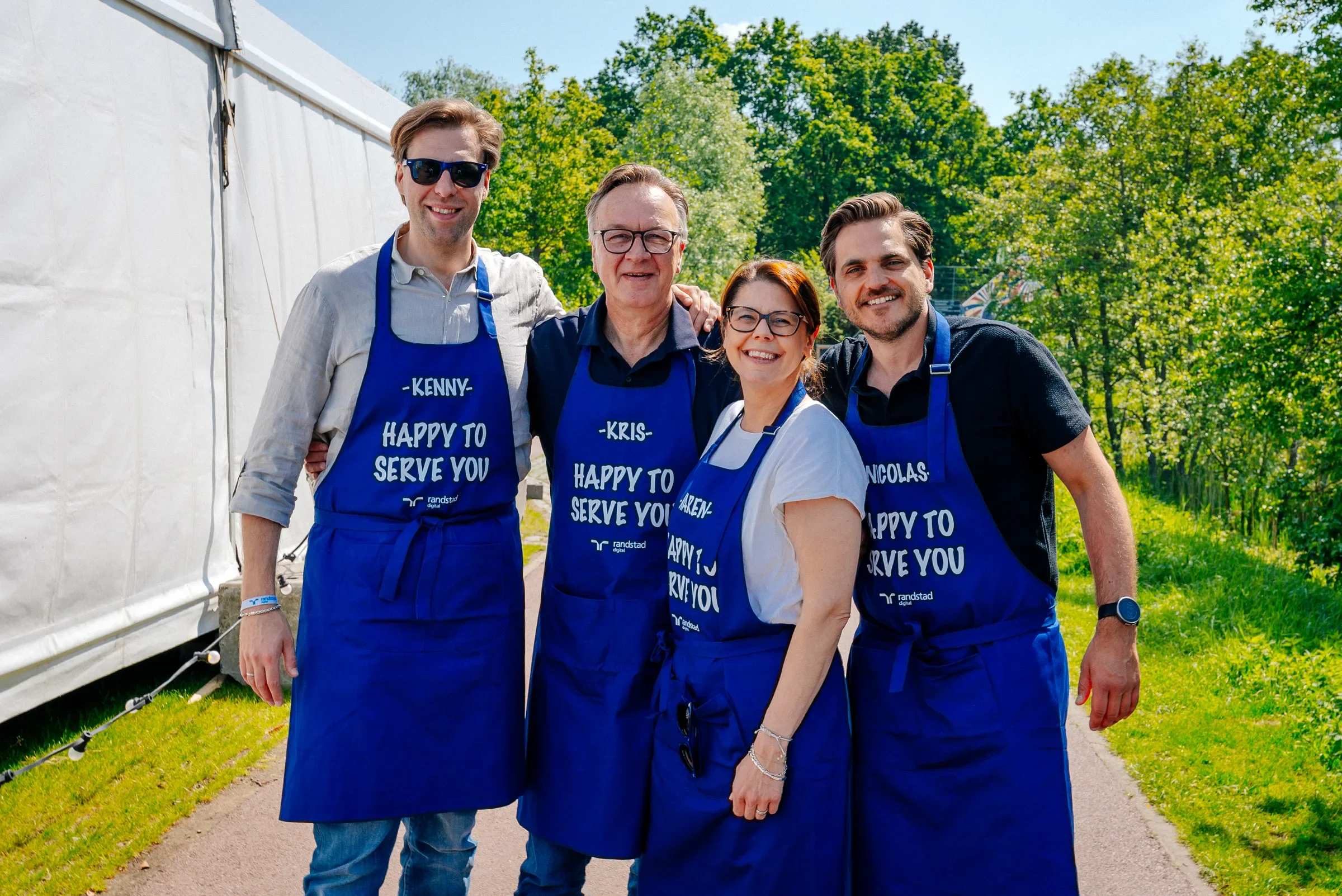 Vijf mensen staan buiten in een groene omgeving, allemaal dragen blauwe aprons met teksten zoals 'Happy to serve you' en namen. Ze lachen en poseren samen voor de foto.
