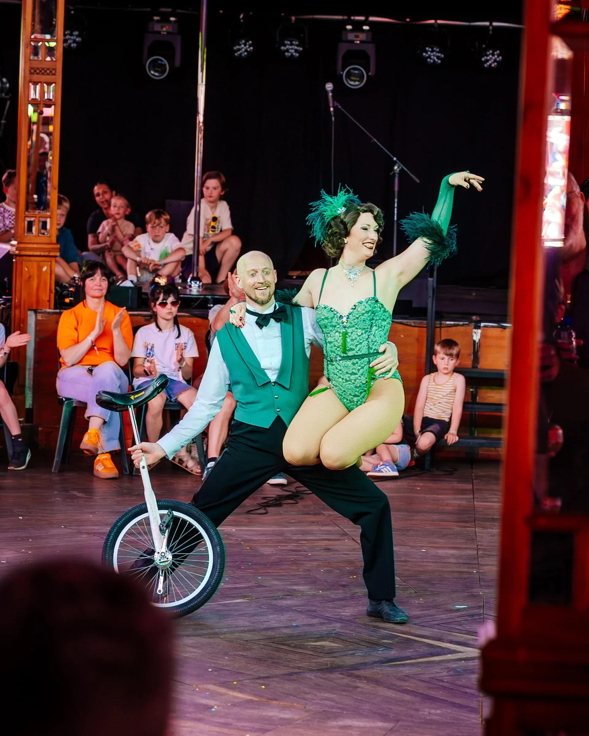Een man en een vrouw in theatrale kostuums voeren een acrobatische act uit op een podium, omringd door een publiek dat uit kinderen en volwassenen bestaat, onder donkere theaterverlichting.