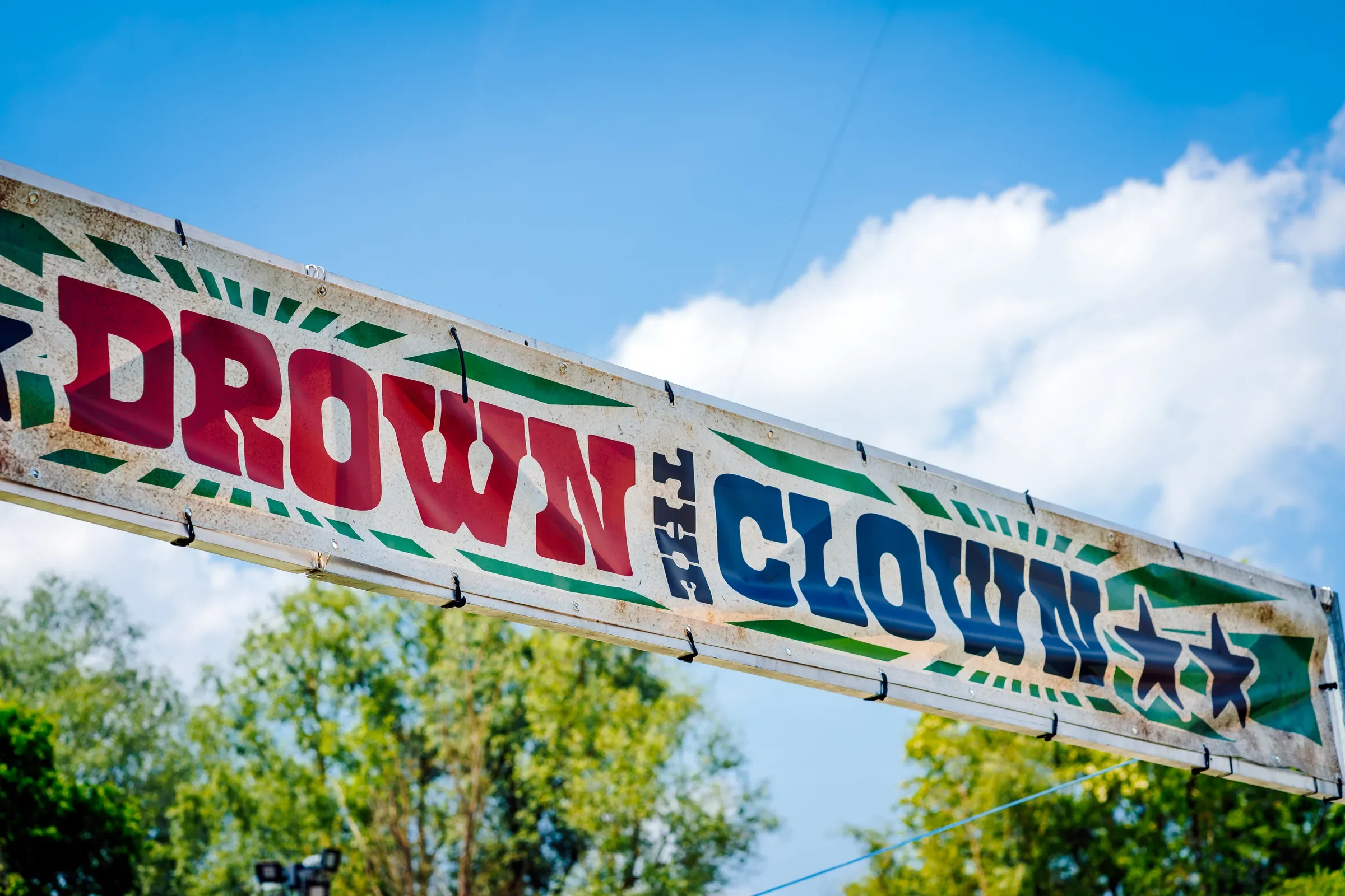 Een kleurrijke reclamebord met de woorden 'CLOSING' en 'CLOS', bevestigd aan een metalen frame onder een blauwe lucht met enkele witte wolken. Op de achtergrond groene bomen.