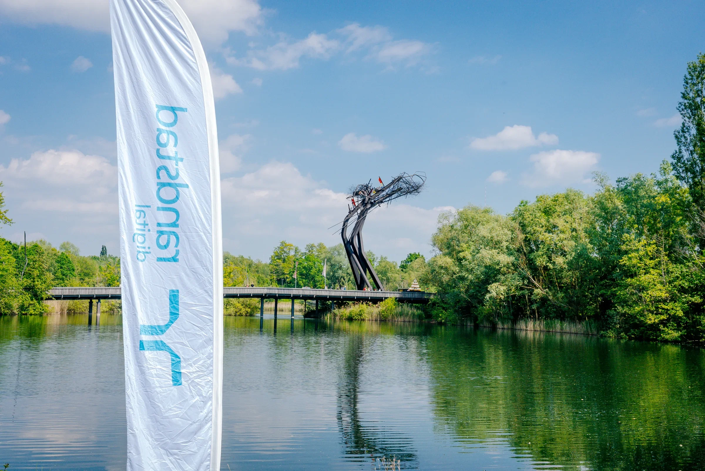 Een vlag met de tekst 'cityseag' naast een rivier, met een kunstwerk van een metalen structuur en een brug op de achtergrond, omringd door groene bomen en een blauwe lucht met wolken.