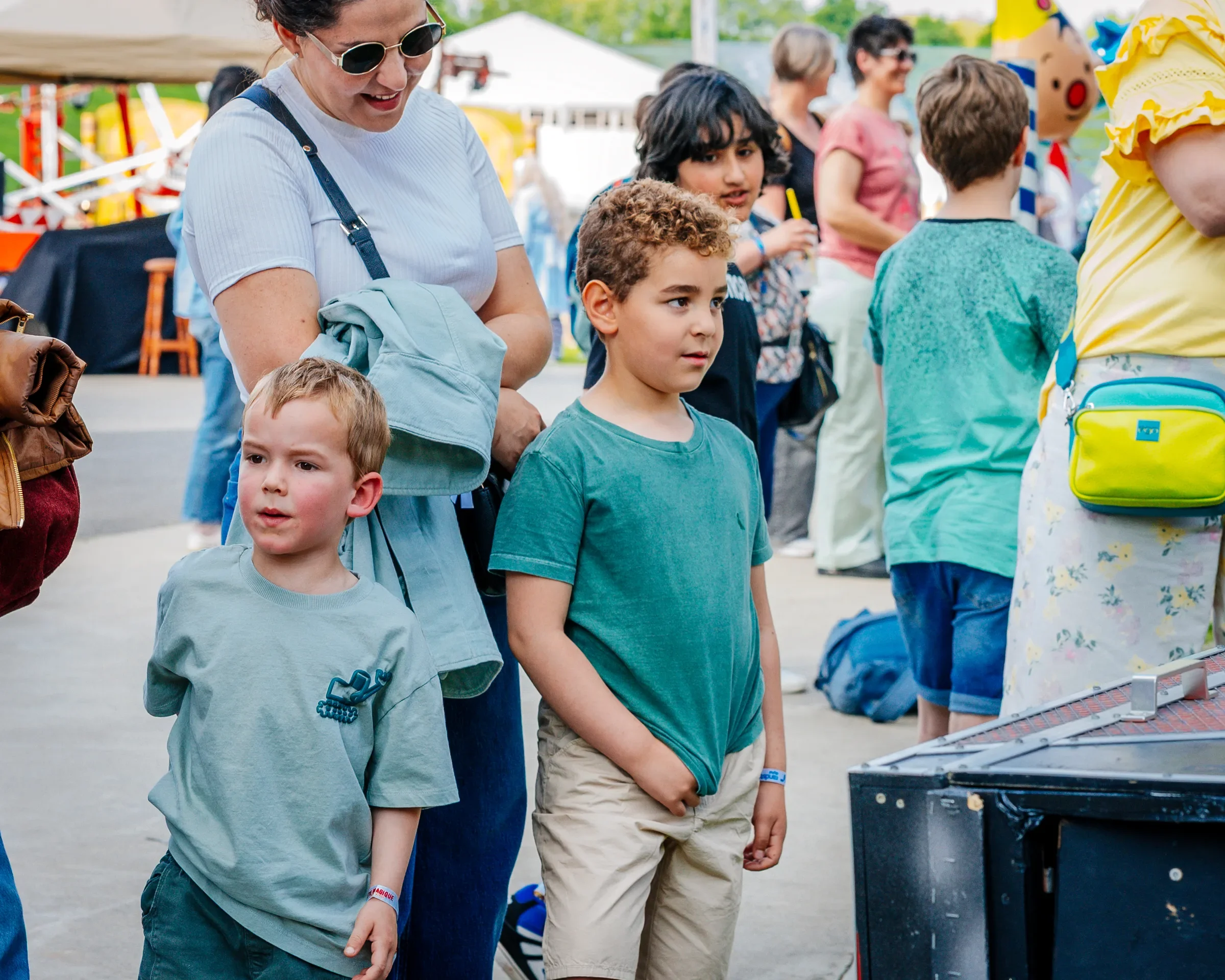 Een groep mensen, voornamelijk kinderen, staan in een lijn en kijken naar een tafel met kleine items op een marktkraam. Een vrouw met donkere haar en een witte shirt kijkt naar beneden terwijl ze naast een jongen met een groene shirt staat. Op de ach