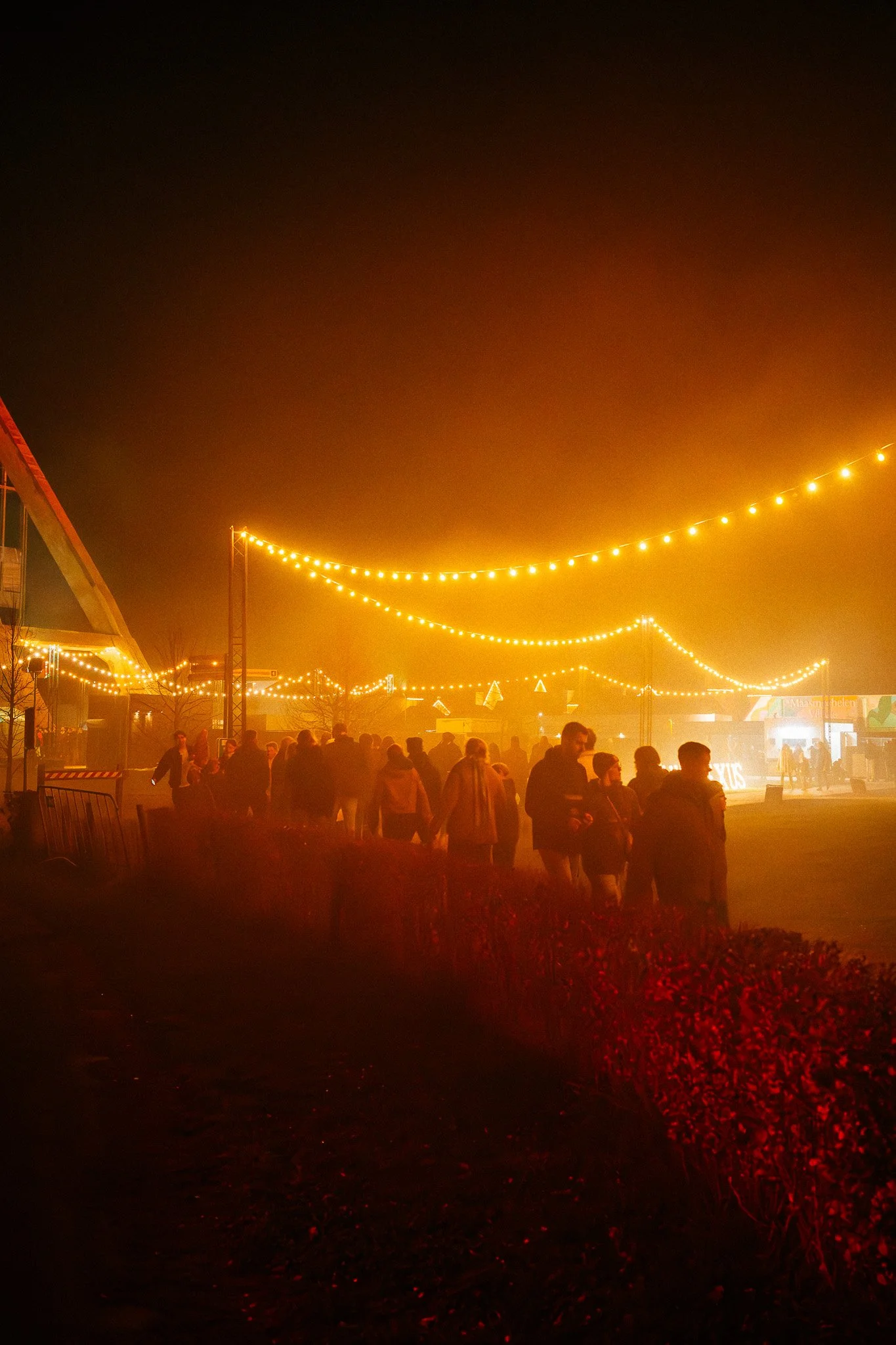 Een groep mensen die 's avonds of 's nachts onder geelverlichte linten op een plein of straat samenkomt.
