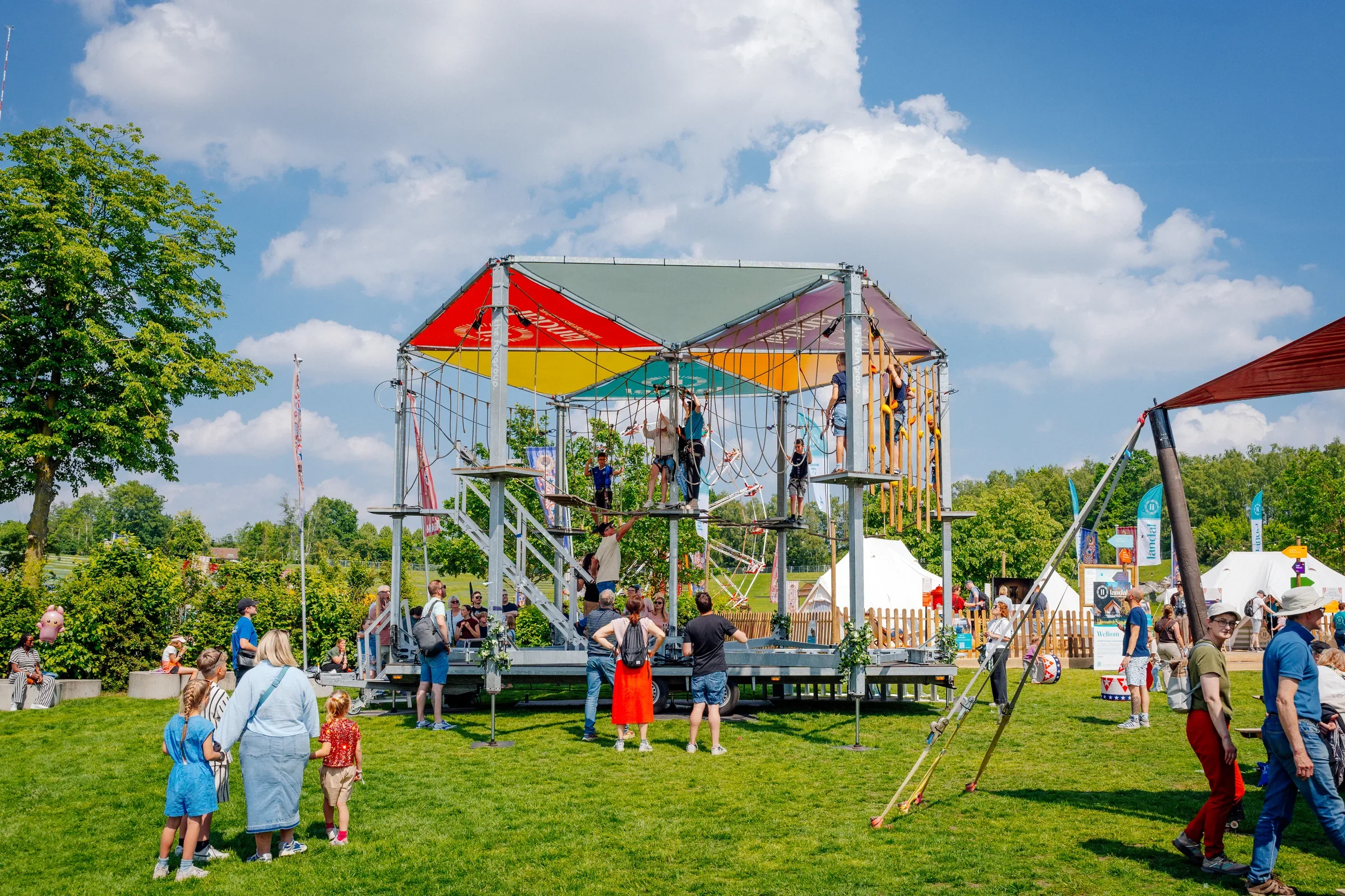 Kinderen en volwassenen genieten van een attractiepark met een klimmuur onder een blauwe hemel met wolken, omringd door groene bomen en witte tenten.