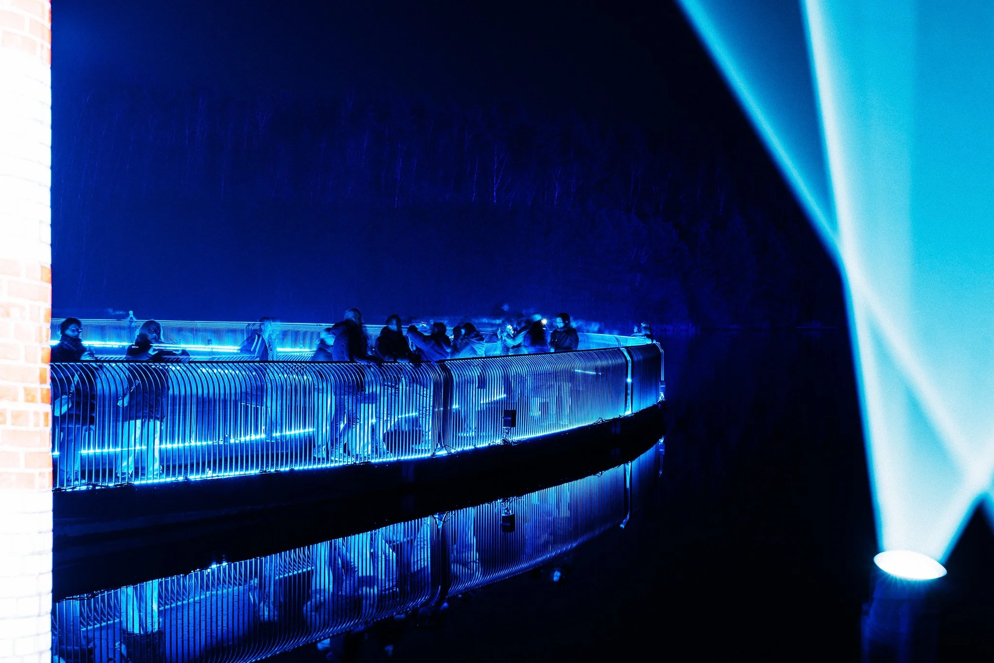 Mensen lopen over een brug met blauwe LED-verlichting in de nacht, omringd door een donkere lucht en bomen op de achtergrond.