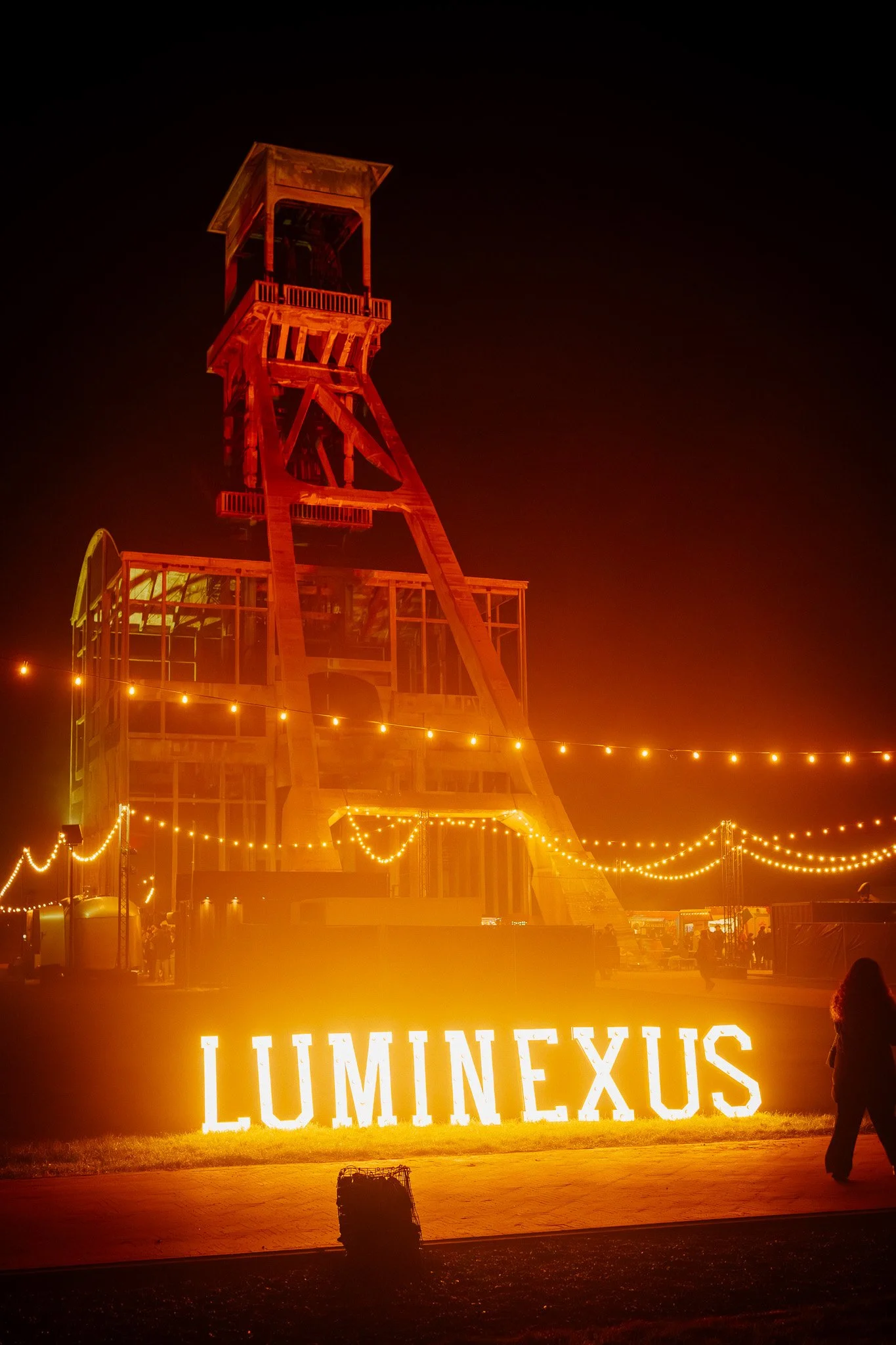 Een verlichte openlucht show of attractie genaamd Lumineux met een grote verlichte tekst op de grond en een houten toren in het midden, omgeven door lichtslingers.