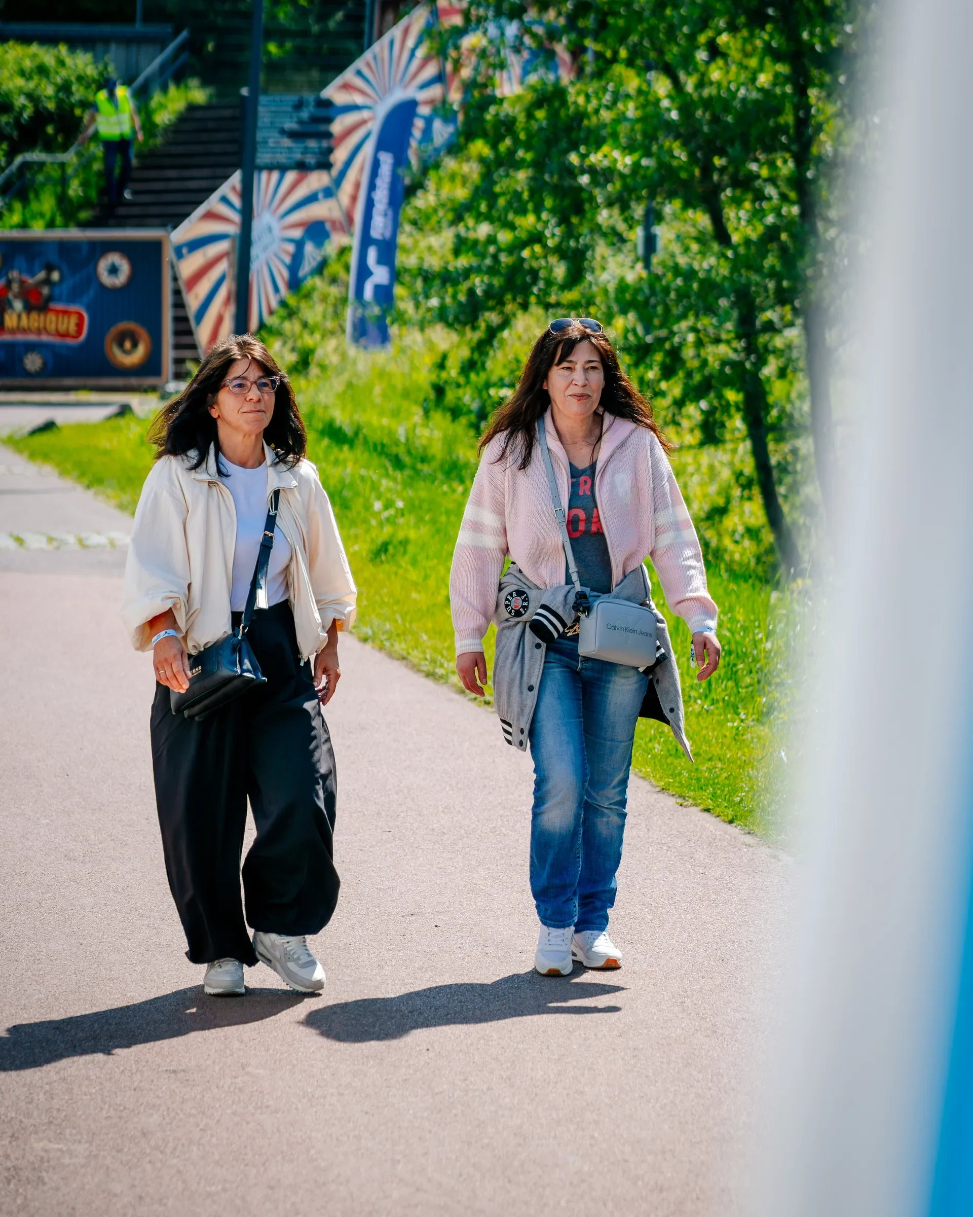Twee vrouwen wandelen op een trottoir in een groene omgeving op een zonnige dag, met kleurrijke parasols en trappen op de achtergrond.