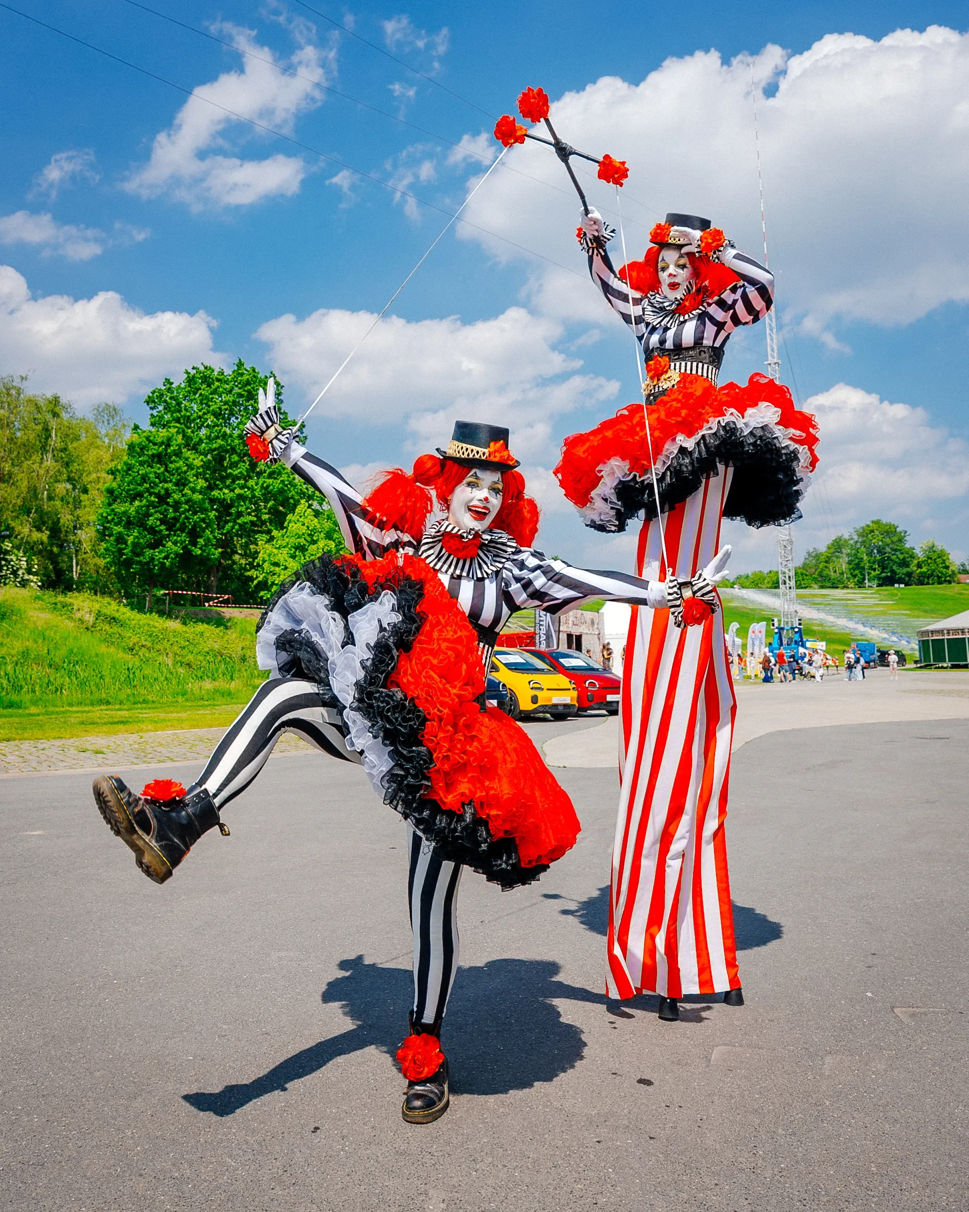 Twee clowns in kleurrijke kostuums, een springt omhoog met één voet op de grond, en de andere in de lucht, terwijl de andere clown naast haar staat met een hand omhoog. De achtergrond toont een open plein met auto's en mensen, groene bomen en een bla