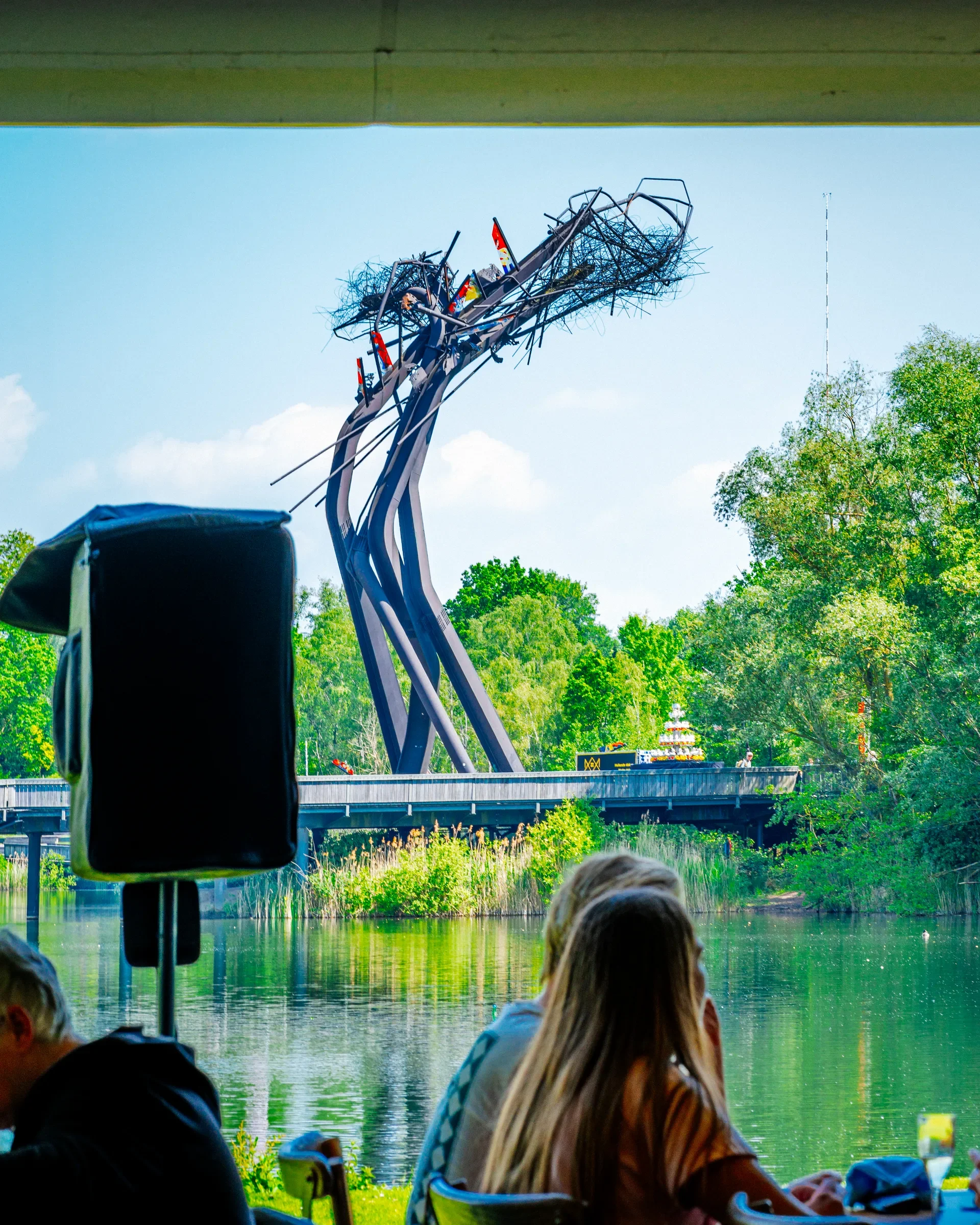 Mensen die in een boot zitten en naar een kunstmatige eendenmoeras kijken met een grote metalen constructie die eruitziet als een nest of takken boven het water, onder een brug.