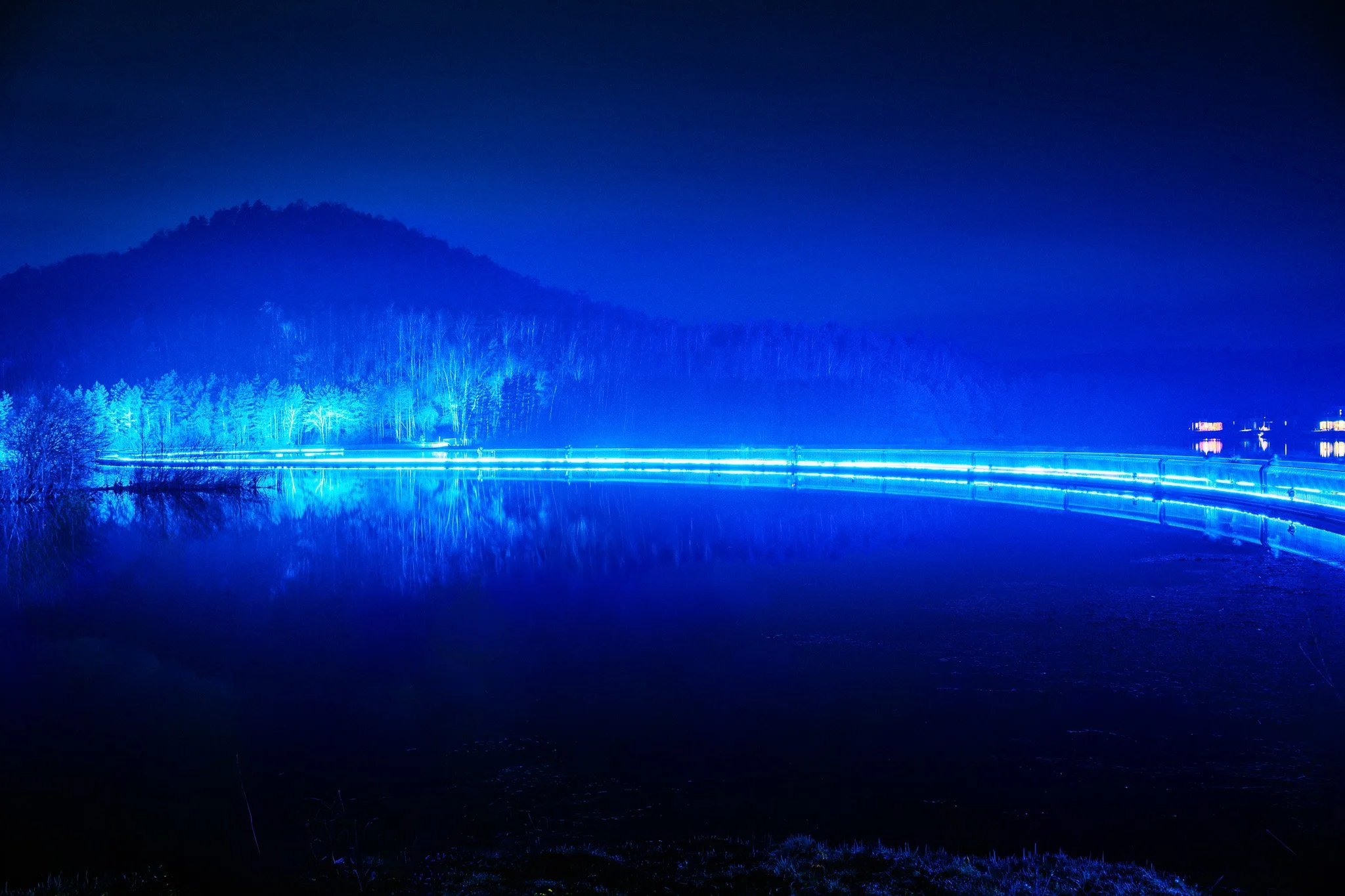 Uitzicht over een rivier met blauwe verlichting, omringd door bomen en heuvels in de verte, 's nachts.