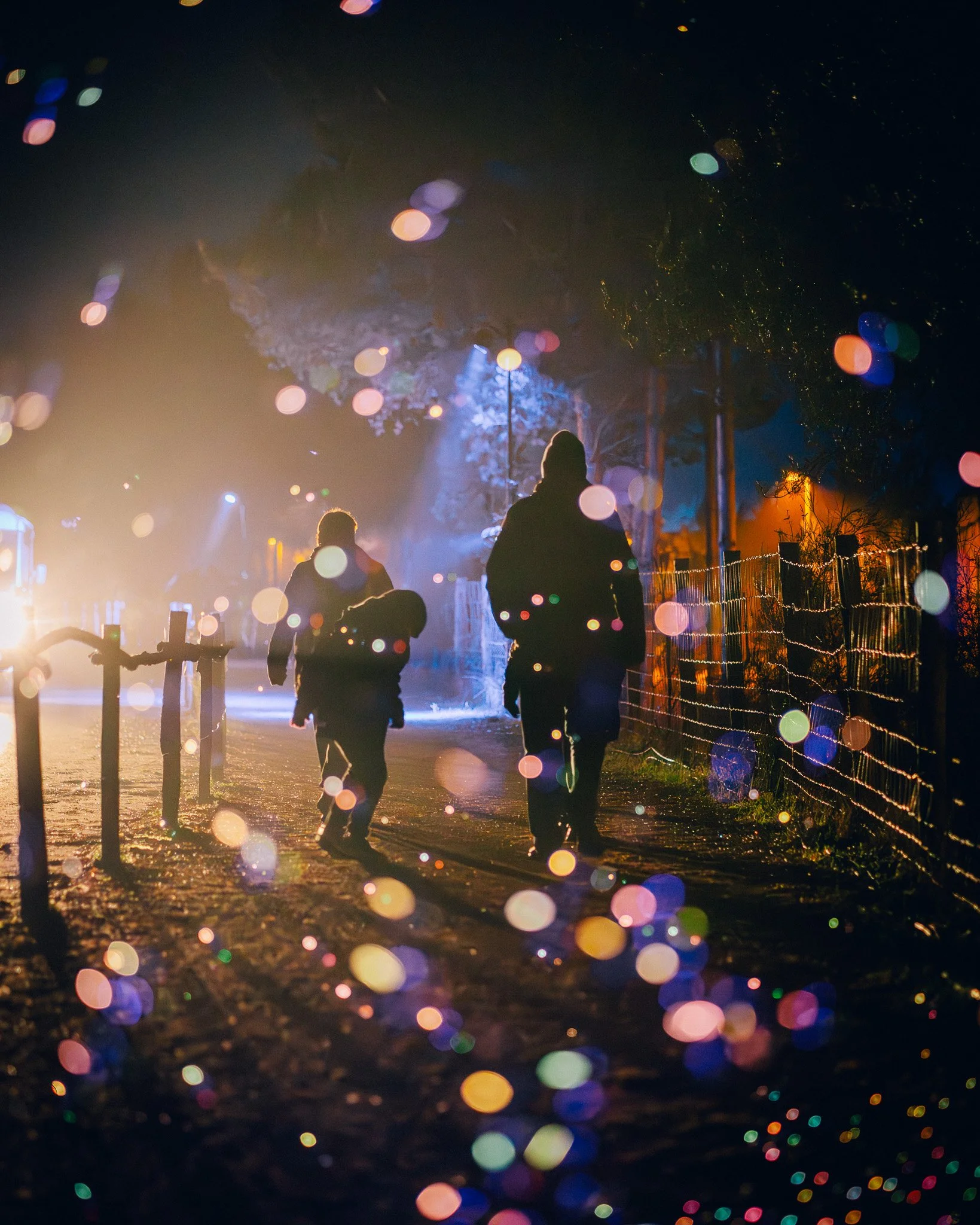 Silhouetten van mensen die wandelen op een verlichte weg 's nachts, met kleurrijke lichtplekken en decoraties in de omgeving.