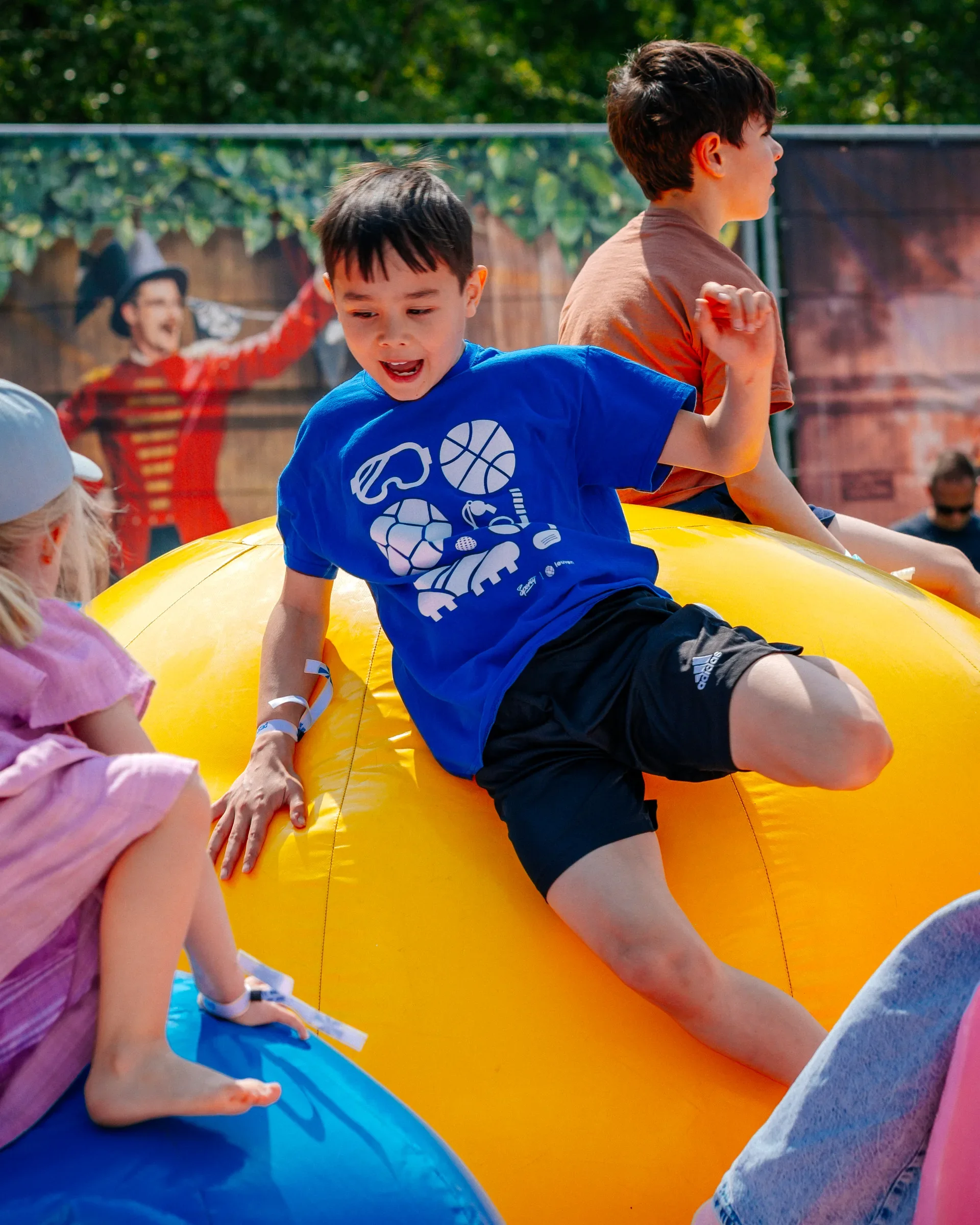 Kinderen die op een opblaasbare ontdekkingstour gaan op een speeltuin met een grote gele en blauwe oppompbare objecten, terwijl een persoon in de achtergrond met een hoed en een wapperend vlagje staat.