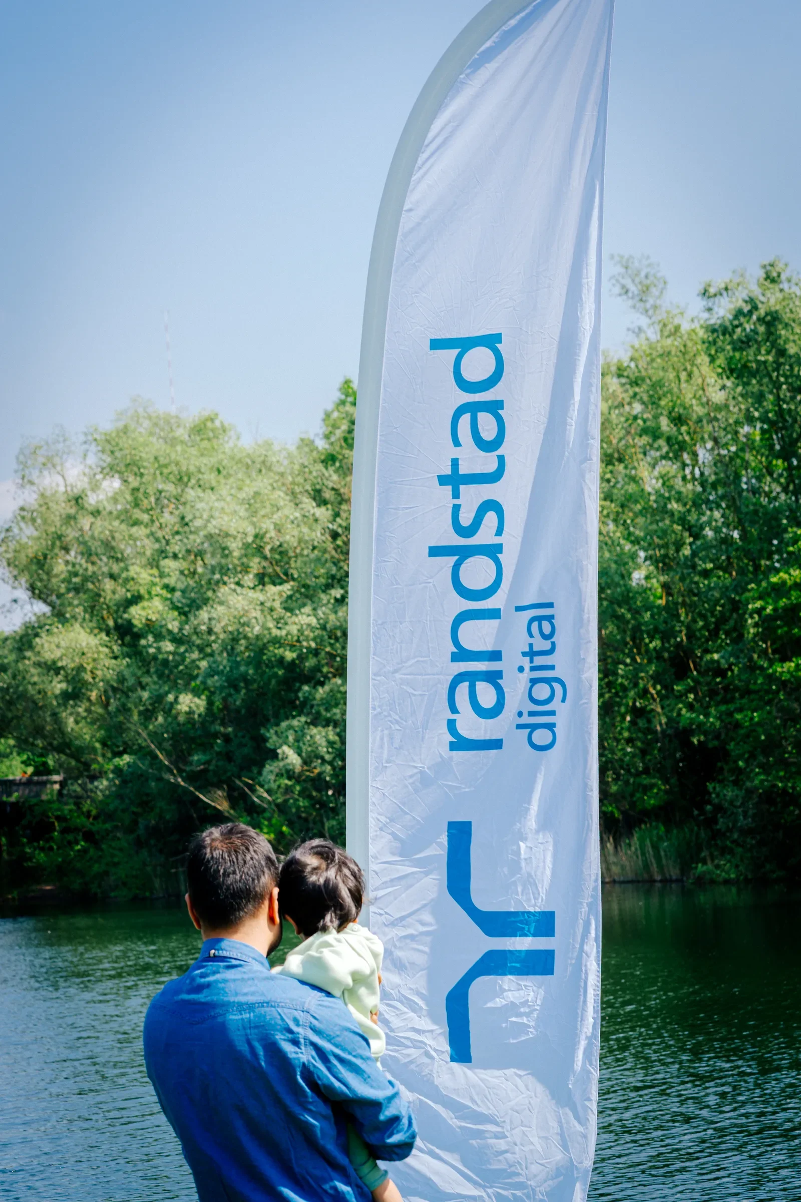 Man met kind bij rivier met vlag van 'arkstad' en bomen op achtergrond.
