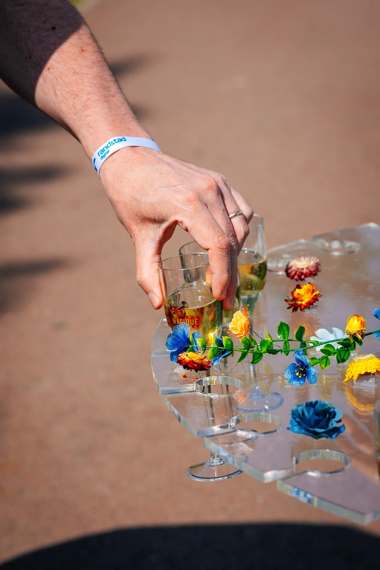 Een hand met een ring en een armband plaatst een glas met een drankje op een doorzichtige tafel die versierd is met kleurrijke bloemen, op een vlakke ondergrond.