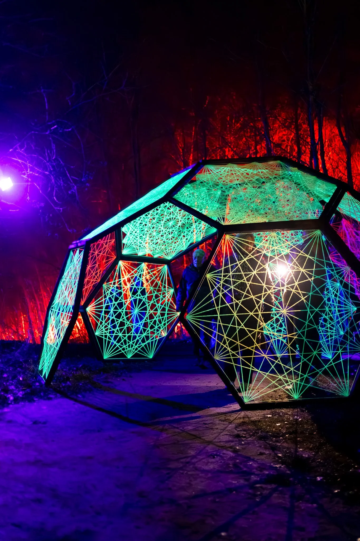 Kleurrijke, verlichte geometrische sculptuur in een donker bos, met wortels die uit de structuur omhoog lijken te komen.