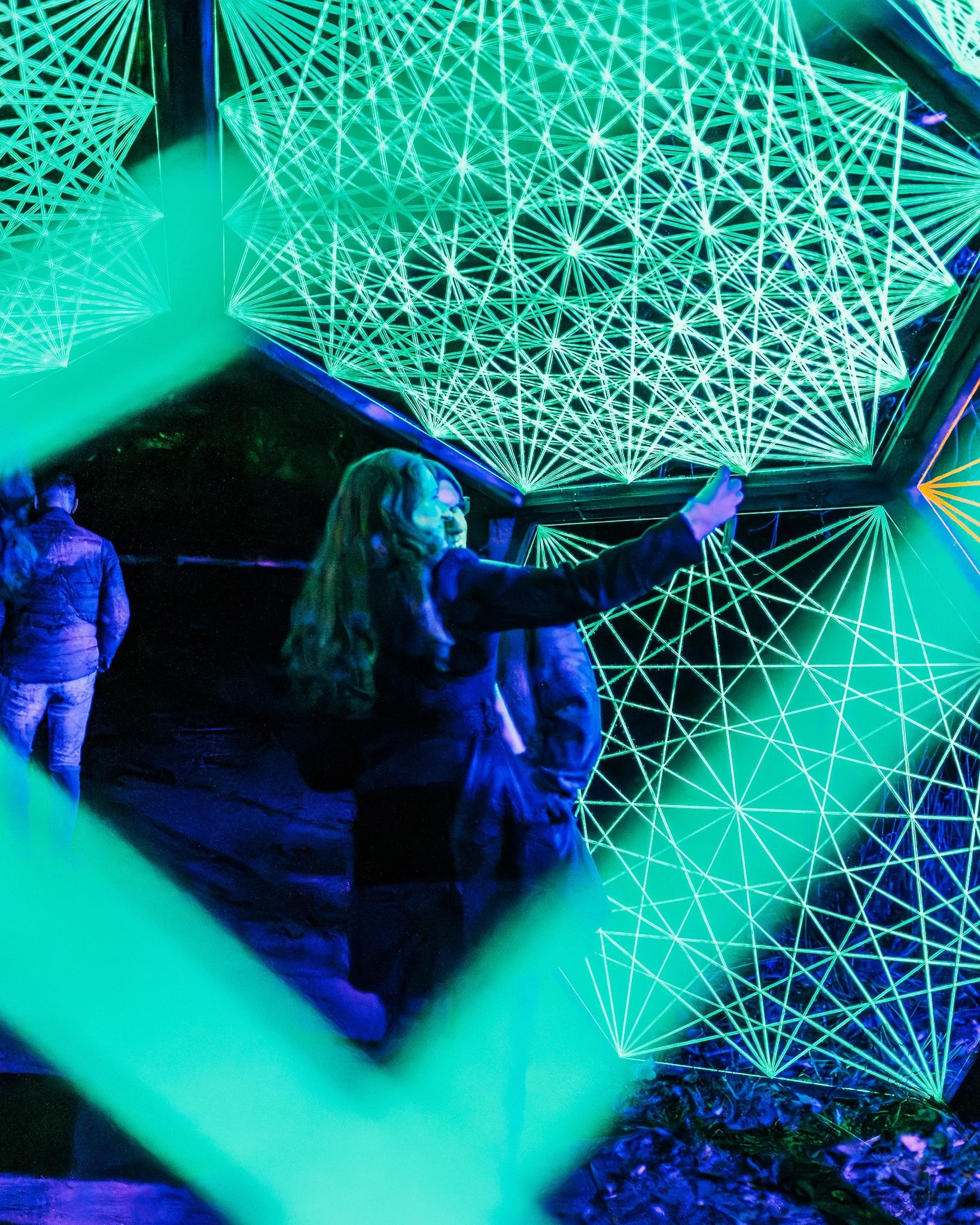 Mensen die naar een neon-lichtinstallatie kijken met geometrische patronen in groen, blauw en paars.