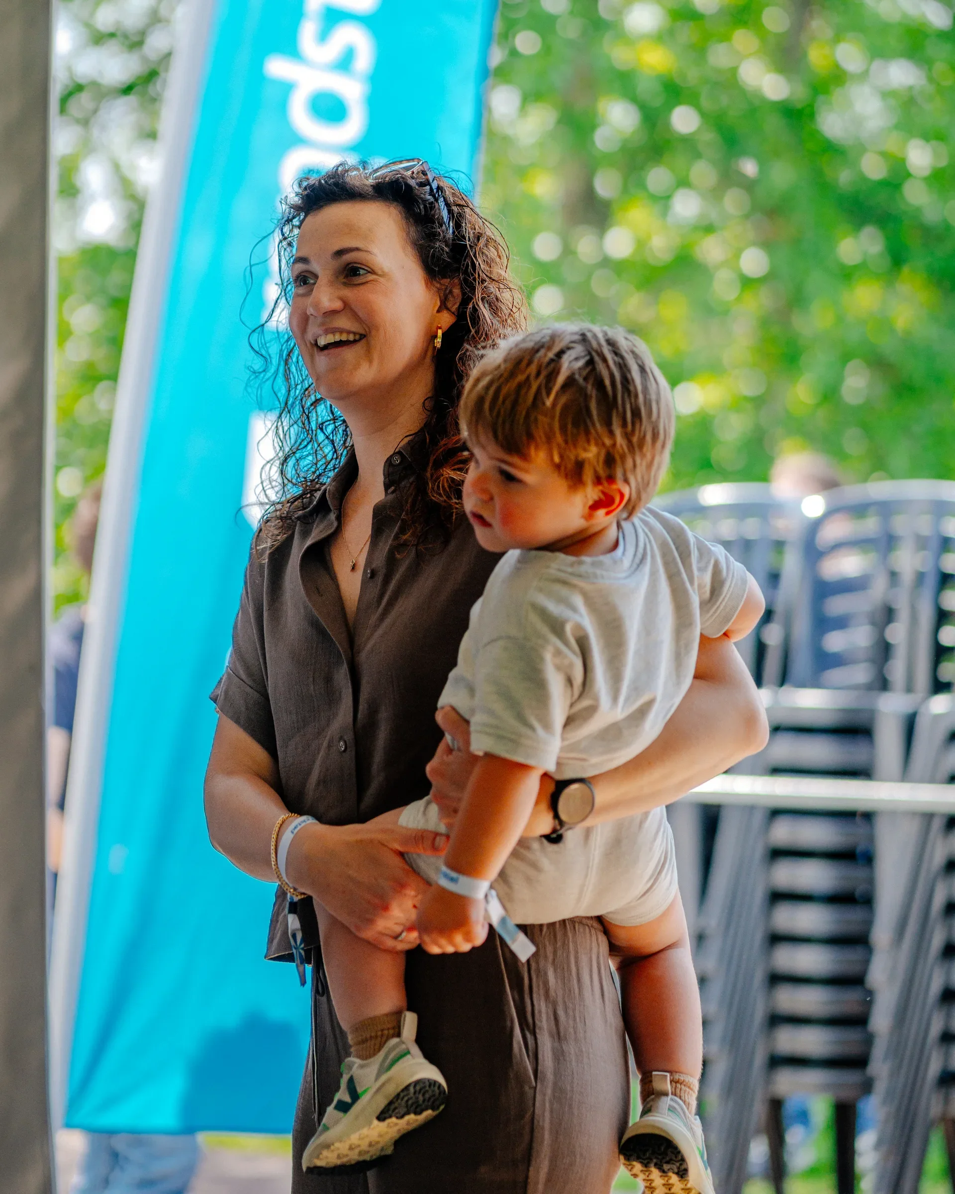 Een vrouw die een jonge jongen draagt, beide lachen. Ze bevinden zich waarschijnlijk in een buitenomgeving met veel groen en er is een blauwe banner op de achtergrond.