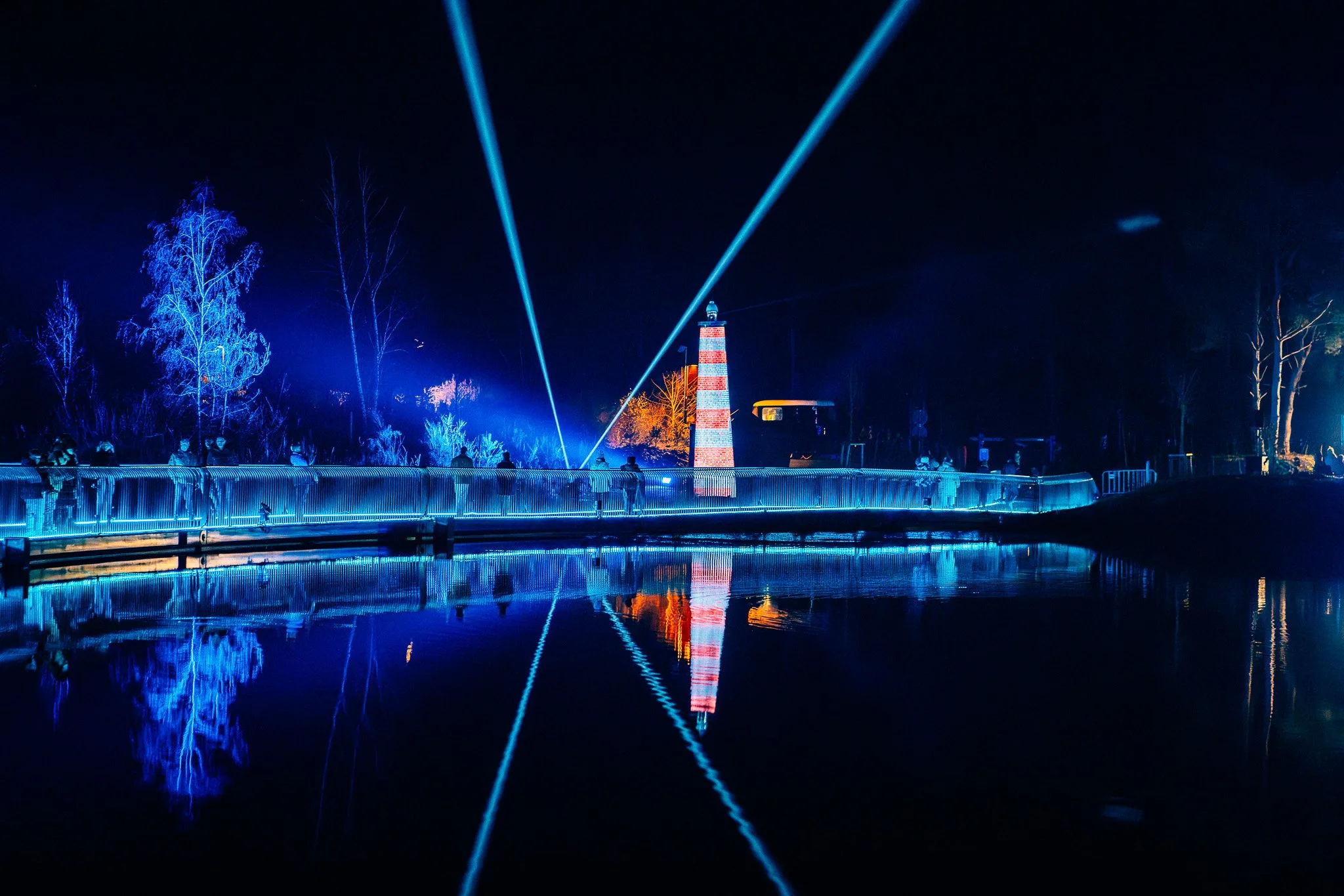 Lichtshows met blauwe en rode laserlijnen boven een waterpartij met gekleurde verlichte bomen op de achtergrond 's nachts.