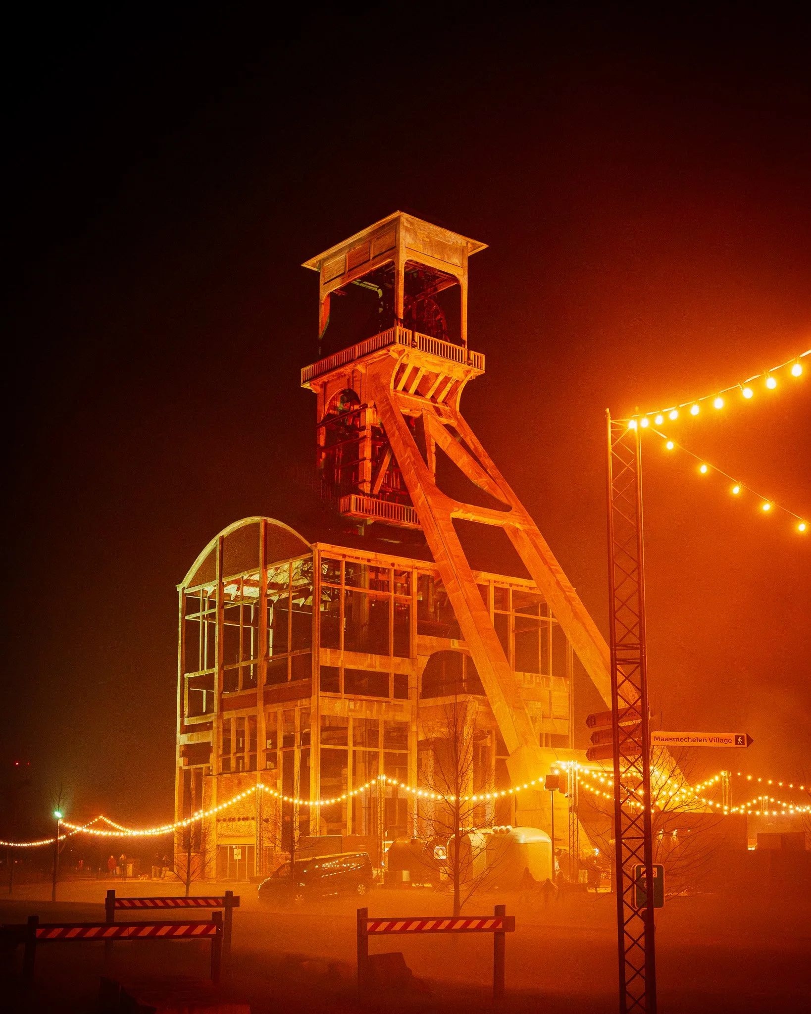 Een houten toren verlicht door rode en gele lichten, omgeven door feestverlichting en mensen op de achtergrond.